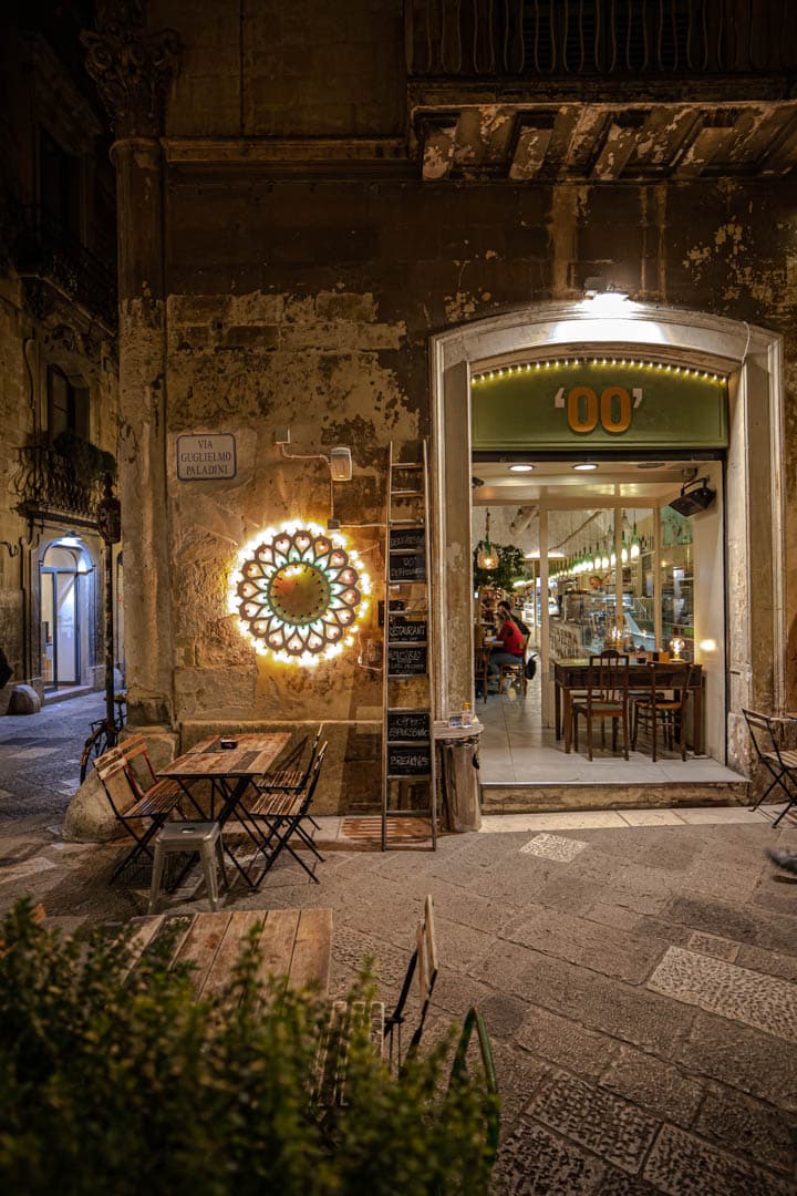 Ein gemütliches Straßencafé mit leeren Tischen sitzt neben einem warm beleuchteten Restauranteingang in einem alten Steingebäude bei Nacht. An der Wand leuchtet eine runde, dekorative Leuchte. © Fotografie Tomas Rodriguez