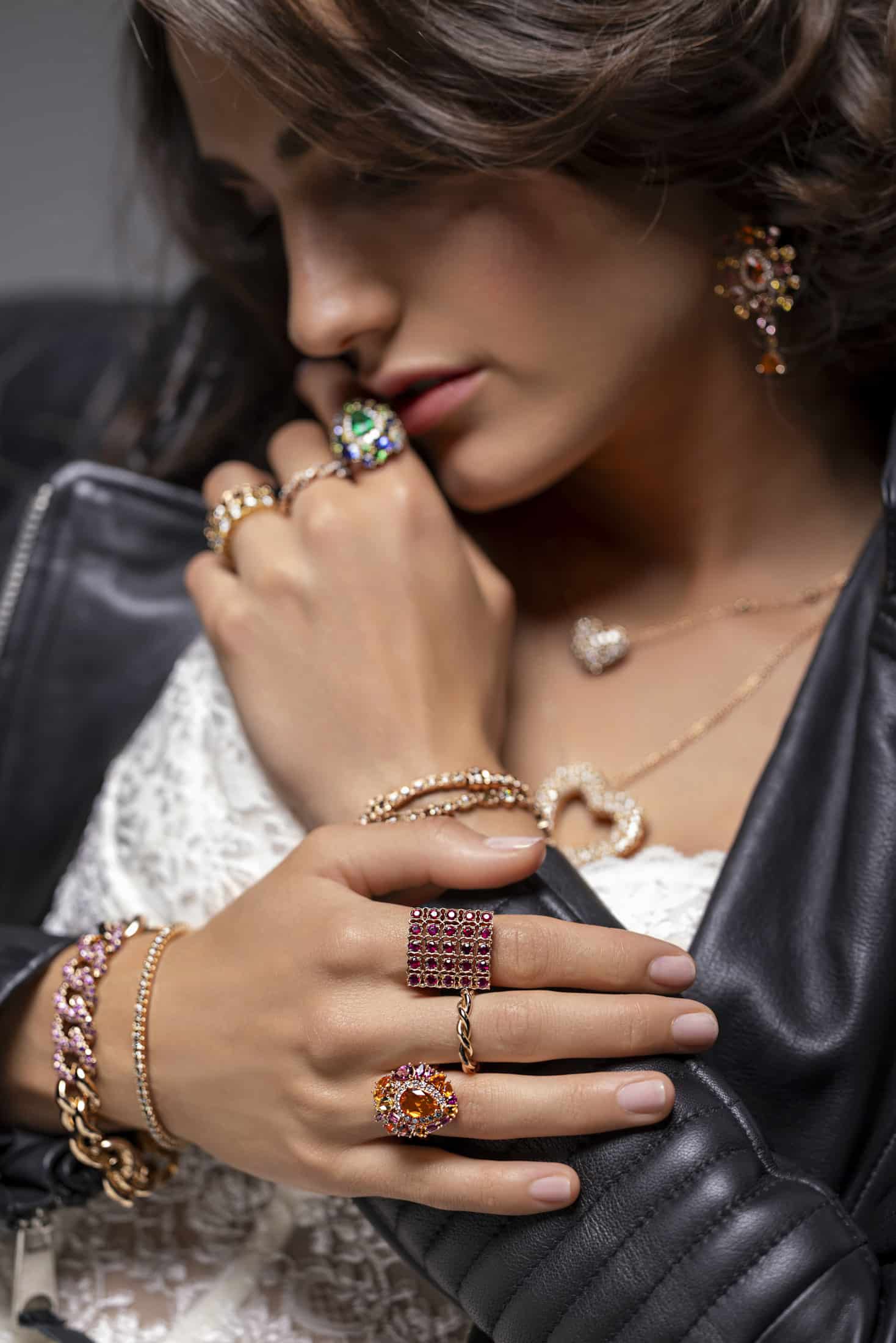 Eine Frau, die eine schwarze Lederjacke und ein Spitzentop trägt, präsentiert mehrere Ringe, Armbänder, eine Halskette und einen Ohrring mit bunten Edelsteinen, hält sich den Kragen und schaut nachdenklich nach unten. © Fotografie Tomas Rodriguez