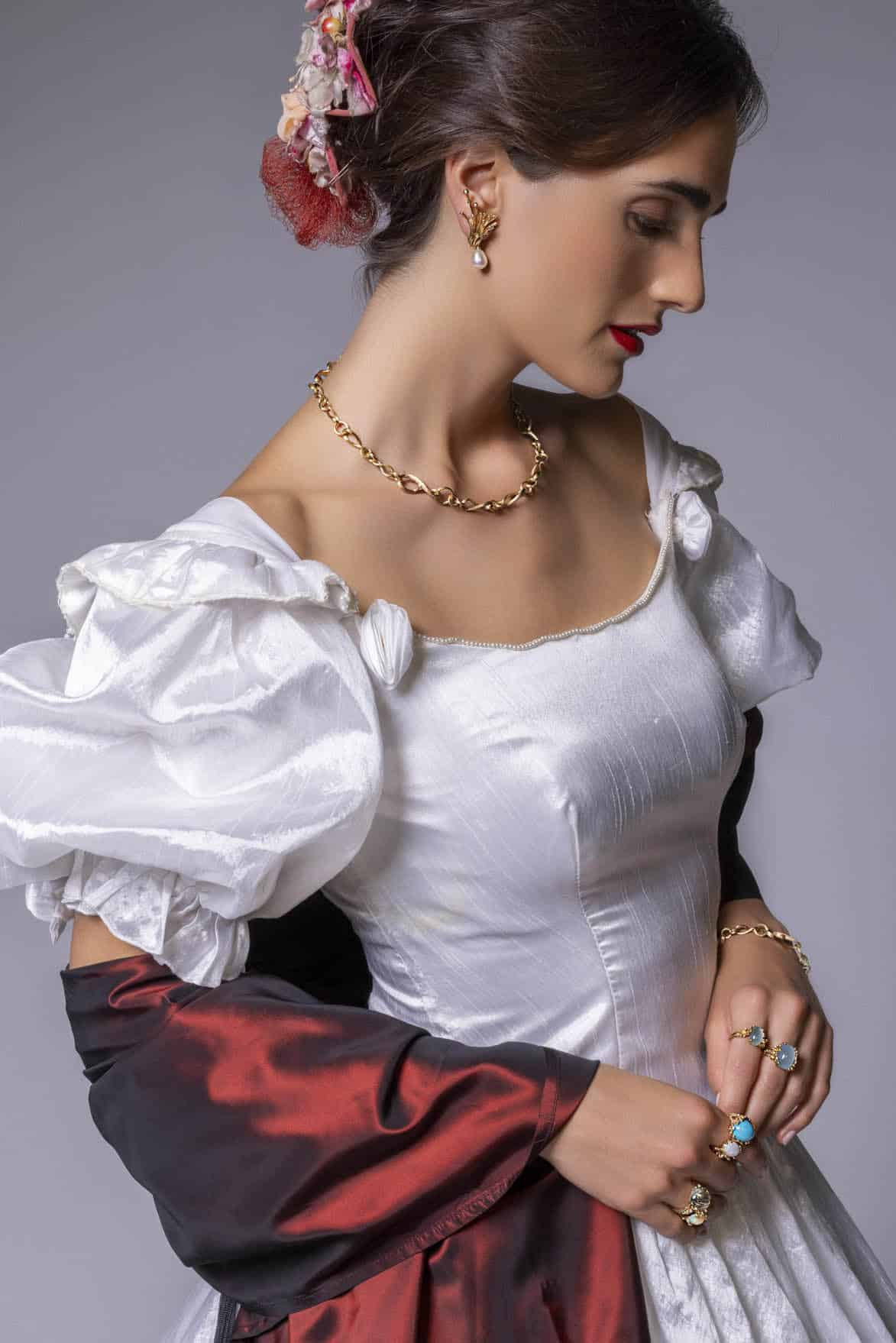 Eine Frau in einem weißen Kleid mit Puffärmeln im Vintage-Stil mit Goldschmuck und bunten Ringen blickt nach unten. Sie trägt ein rot-schwarzes Tuch und hat Blumen im Haar, die zu einer eleganten Hochsteckfrisur frisiert sind. © Fotografie Tomas Rodriguez
