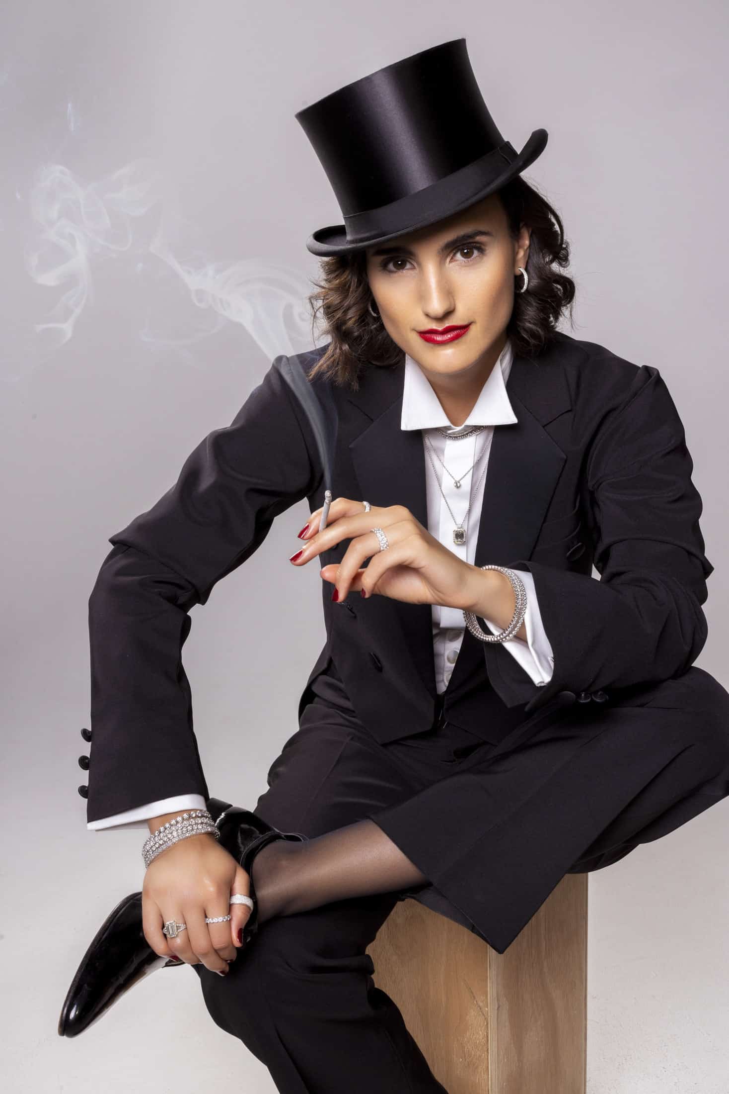 Eine Person, die einen schwarzen Smoking, ein weißes Hemd und einen Zylinder trägt, sitzt auf einer Holzkiste und hält eine Zigarette in der Hand, aus der Rauch aufsteigt. Sie trägt kurzes dunkles Haar, roten Lippenstift und Schmuck und nimmt eine selbstbewusste und stilvolle Pose ein. © Fotografie Tomas Rodriguez