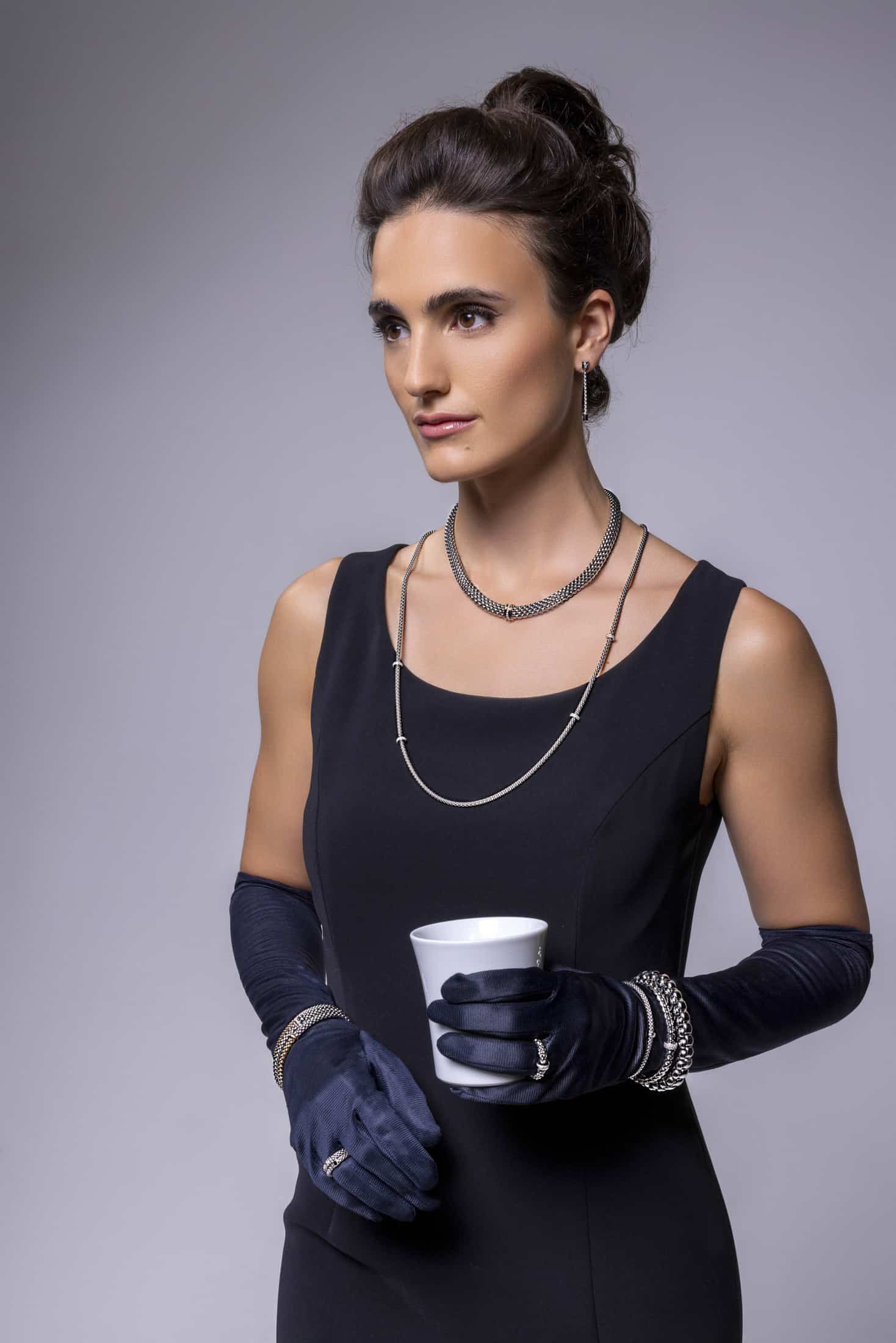 Eine Frau in einem eleganten schwarzen Kleid und langen schwarzen Handschuhen hält eine weiße Tasse. Sie trägt mehrlagige Ketten, Armbänder, Ringe und Ohrstecker, ihr Haar ist hochgesteckt, vor einem schlichten grauen Hintergrund. © Fotografie Tomas Rodriguez