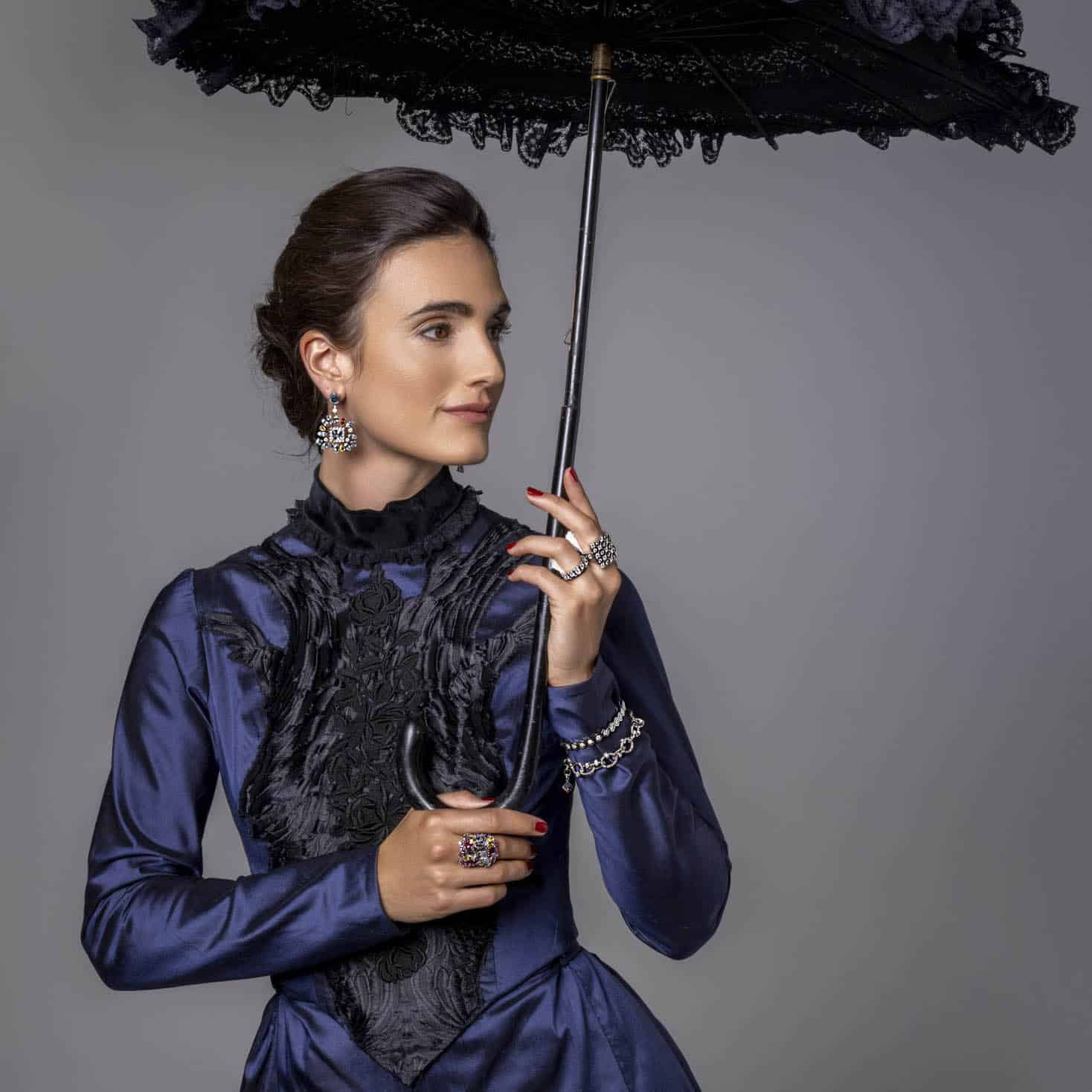 Eine Frau in einem dunkelblauen Kleid im viktorianischen Stil hält einen Sonnenschirm aus schwarzer Spitze. Sie trägt verzierten Schmuck, darunter Ohrringe, Ringe und ein Armband, und hat ihr dunkles Haar zu einer eleganten Hochsteckfrisur frisiert. © Fotografie Tomas Rodriguez