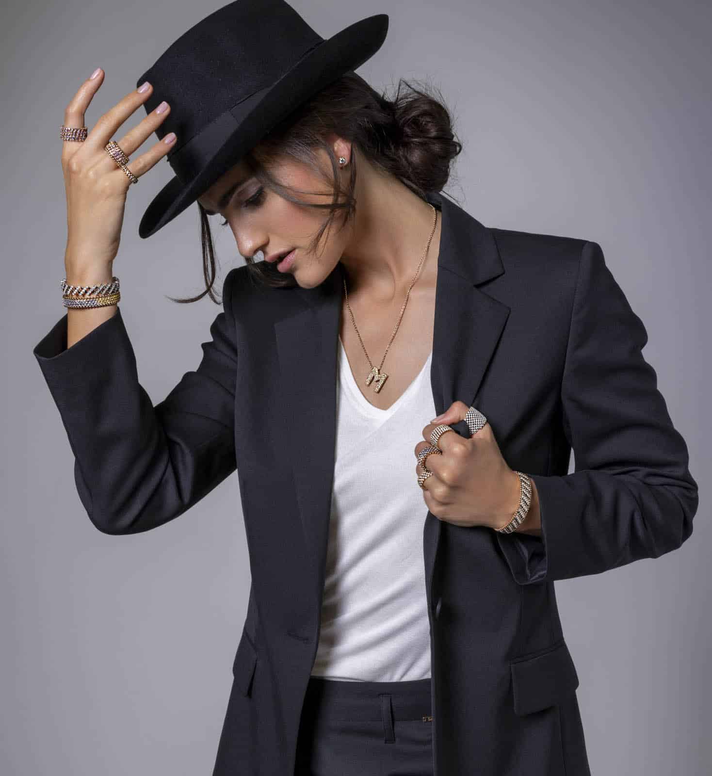 Eine Frau in einem schwarzen Anzug und weißem Hemd neigt ihren schwarzen Hut nach unten und blickt nach unten. Sie trägt mehrere Ringe, ein Armband und eine Halskette und hat ihr Haar zu einem lockeren Dutt frisiert. Der Hintergrund ist einfarbig grau. © Fotografie Tomas Rodriguez