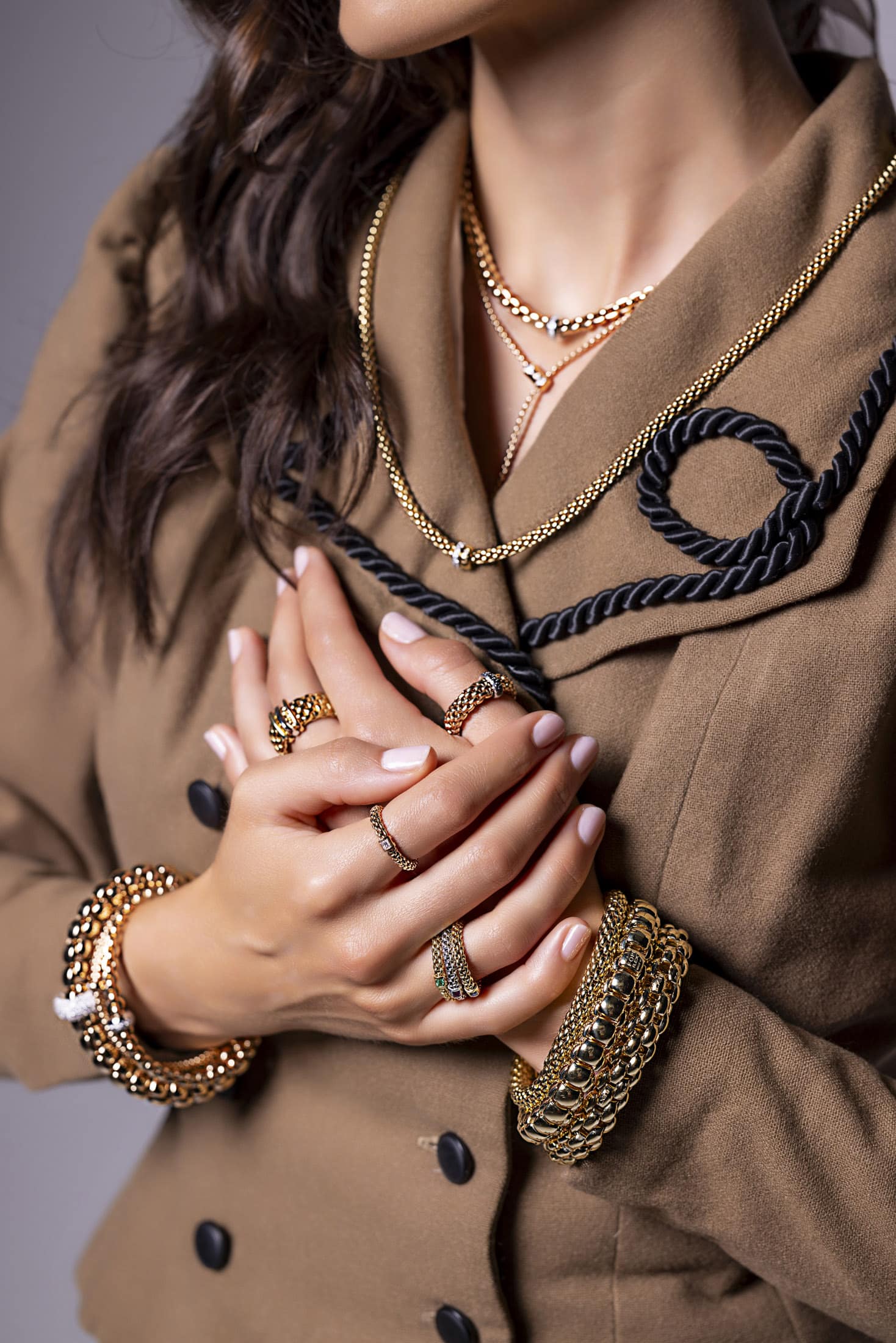 Eine Frau, die einen braunen Mantel mit schwarzem Besatz, mehrere goldene Halsketten, mehrere goldene Armbänder und Ringe an beiden Händen trägt und die Hände vor der Brust verschränkt. © Fotografie Tomas Rodriguez