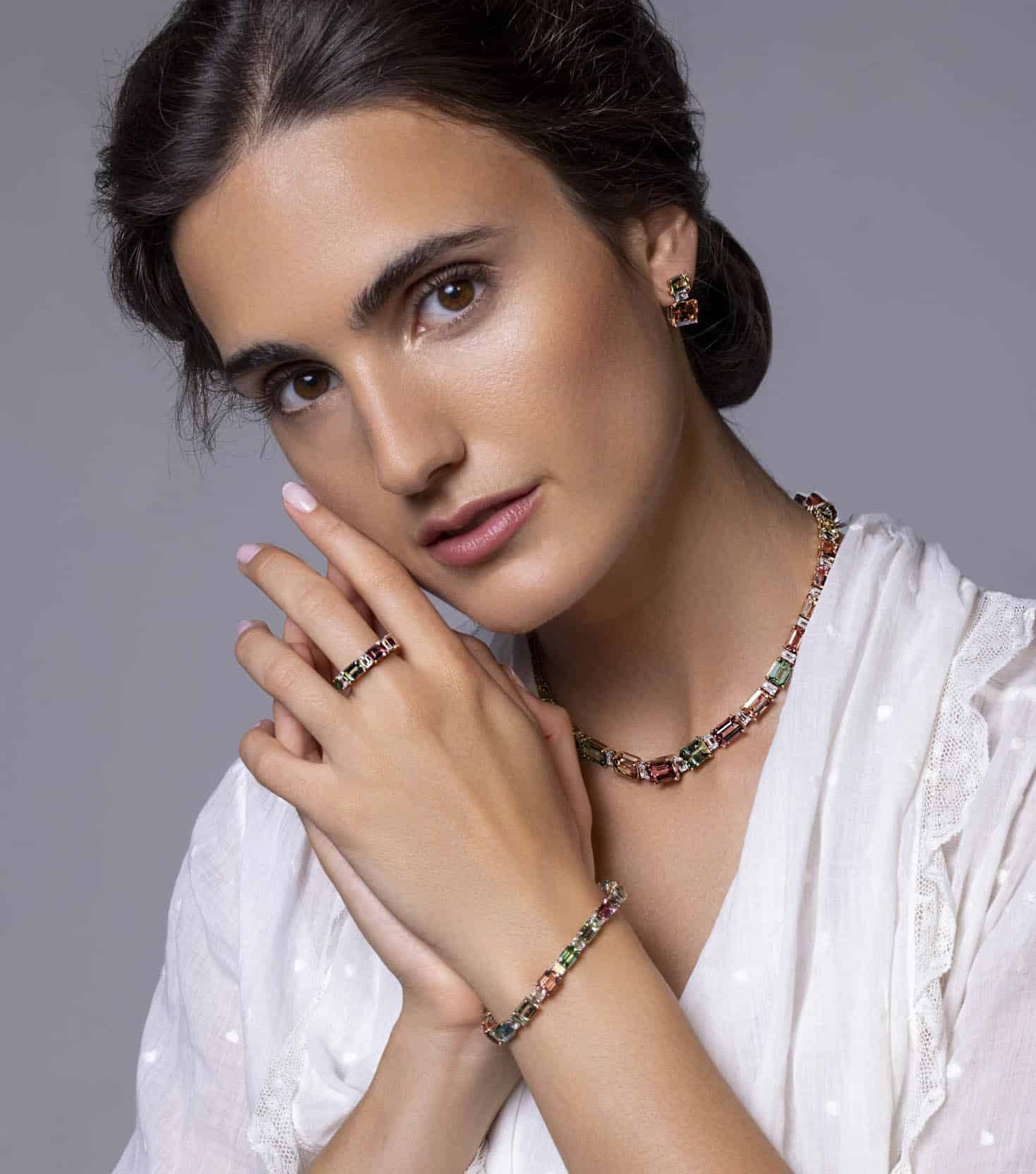 Eine Frau in einer weißen Bluse posiert mit der Hand in der Nähe ihres Gesichts und trägt eine bunte Edelsteinkette, Ohrringe, ein Armband und einen Ring, die alle das gleiche Design haben. Sie trägt ein natürliches Make-up und dunkelbraunes, nach hinten gezogenes Haar. © Fotografie Tomas Rodriguez