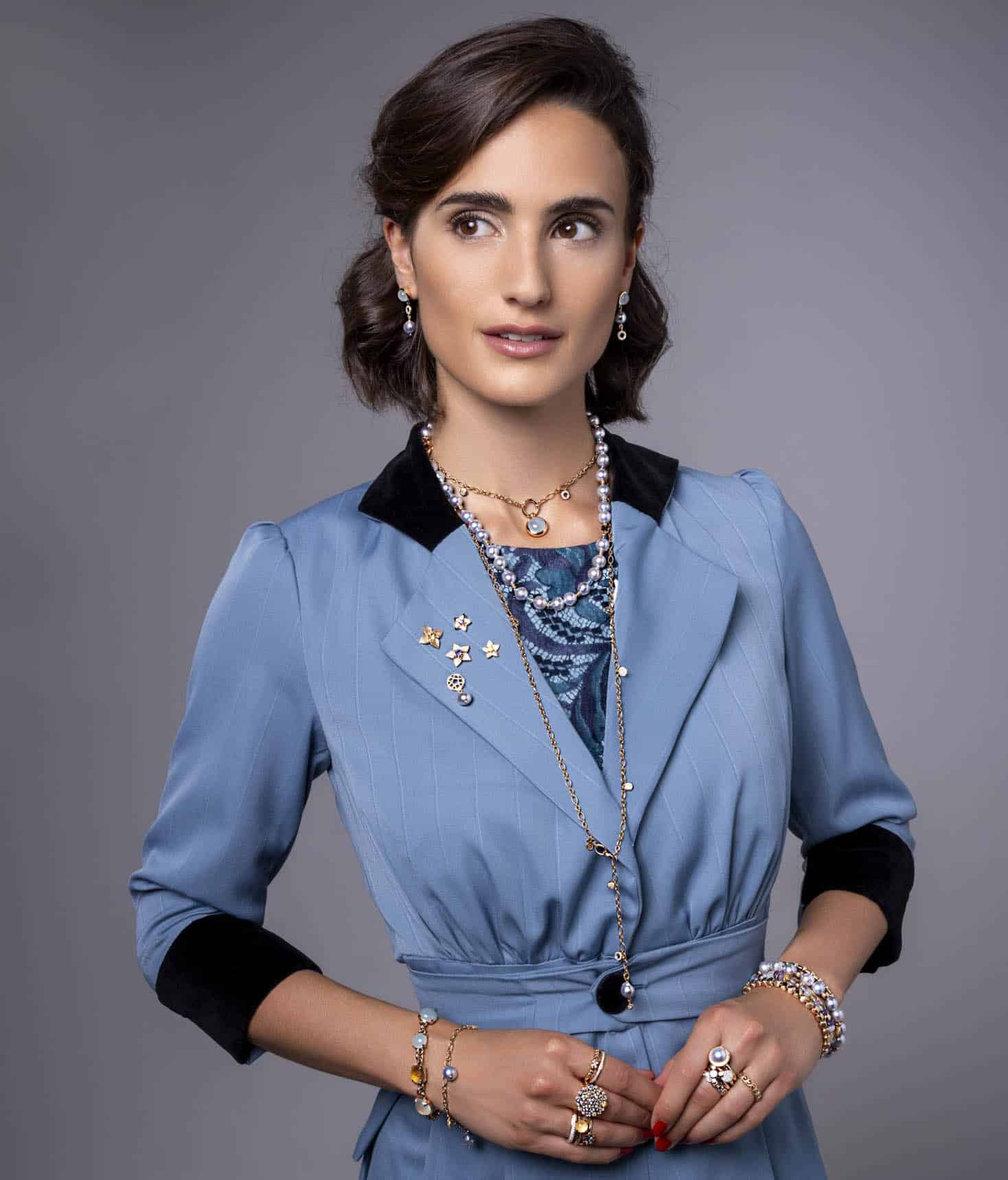 Eine Frau mit kurzen braunen Haaren trägt ein blaues Kleid mit schwarzem Kragen und Manschetten, mehrlagige Halsketten, Ringe, Armbänder und Broschen. Sie blickt leicht zur Seite und steht vor einem schlichten grauen Hintergrund. © Fotografie Tomas Rodriguez