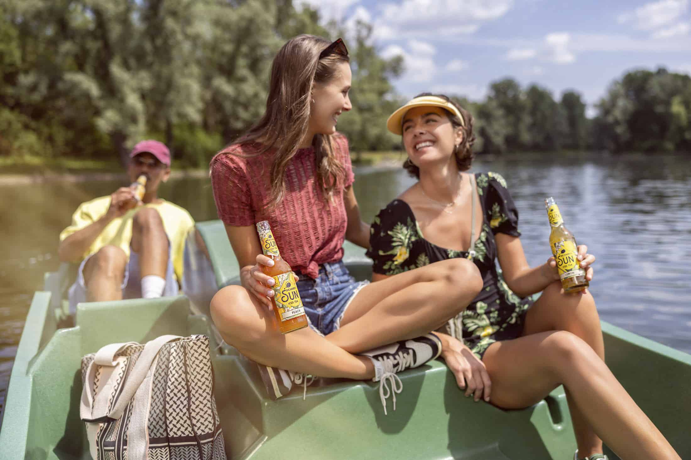 Zwei junge Frauen sitzen lächelnd auf einem Boot, jede hält ein Getränk in der Hand. Ein Mann im Hintergrund trinkt ebenfalls. Die Gruppe befindet sich auf einem ruhigen Fluss mit Bäumen und blauem Himmel im Hintergrund. © Fotografie Tomas Rodriguez