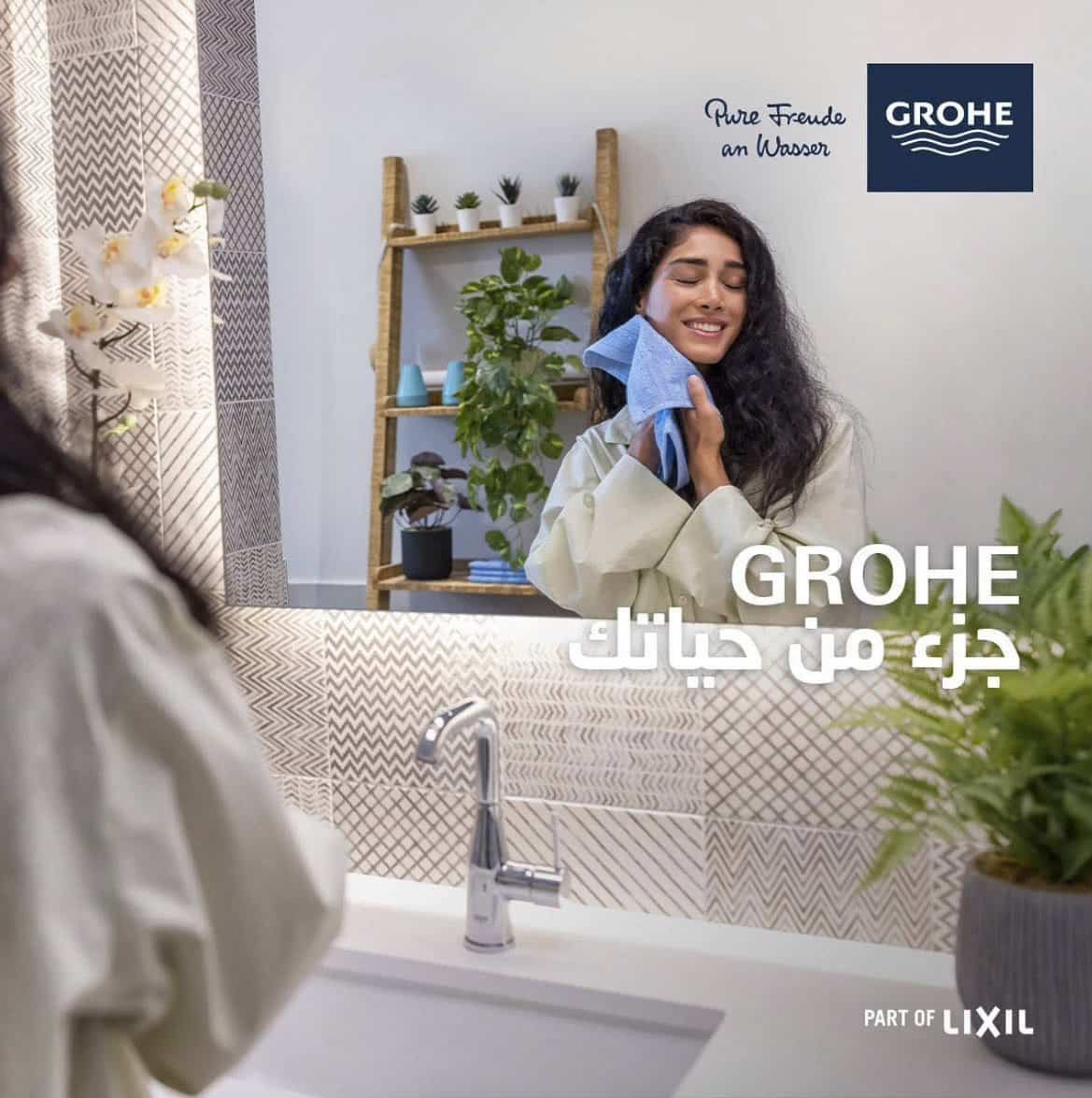 Eine Frau im Bademantel lächelt und trocknet ihr Gesicht mit einem Handtuch in einem modernen Badezimmer ab. Das GROHE Logo und der arabische Text sind abgebildet, im Hintergrund sind Pflanzen und ein Spiegel zu sehen. © Fotografie Tomas Rodriguez