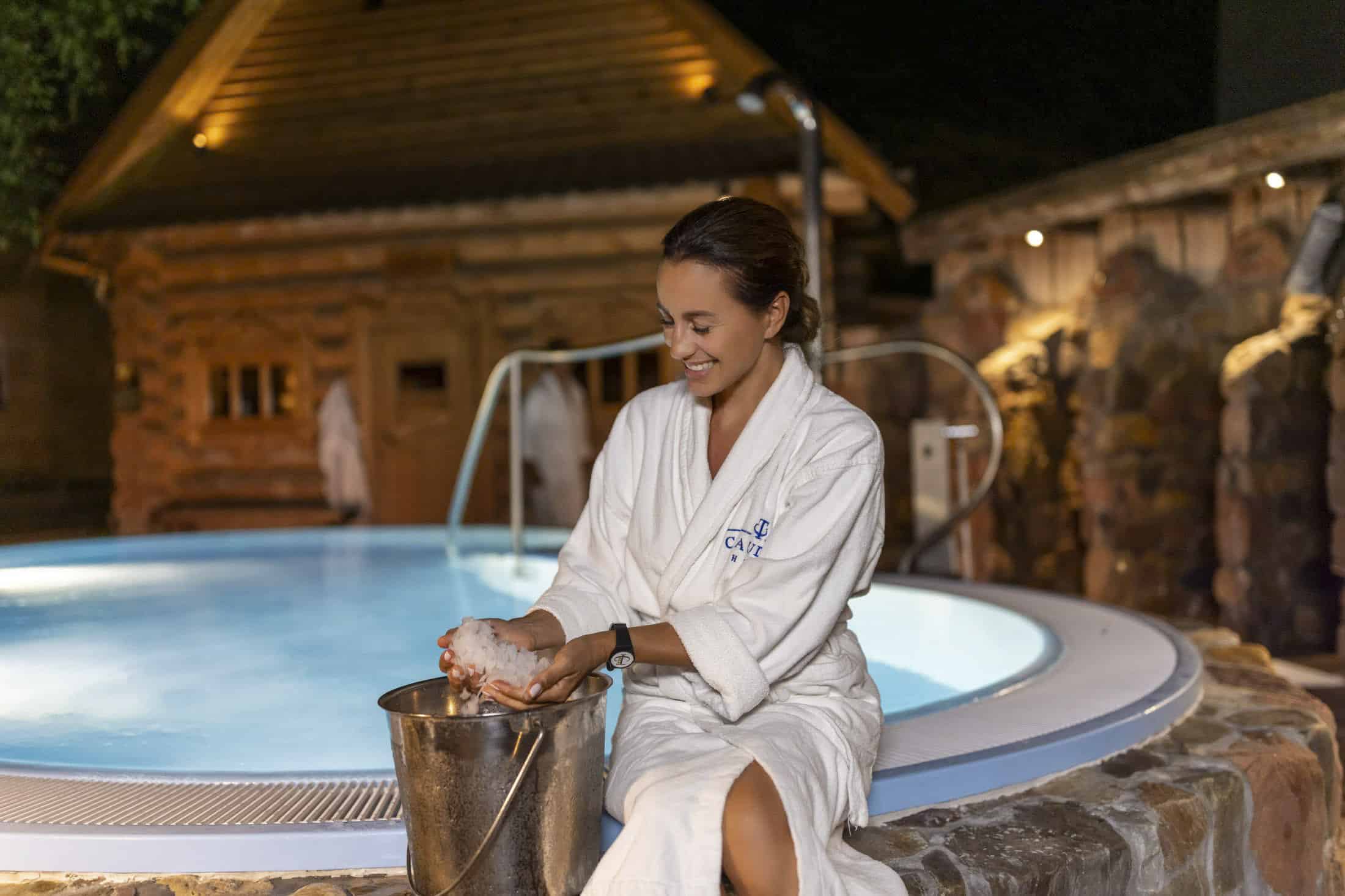 Eine Frau in einem weißen Gewand sitzt nachts an einem Whirlpool im Freien und lächelt, während sie über einem Metalleimer Schnee in ihren Händen hält. Im Hintergrund sind ein Holzgebäude und eine warme, stimmungsvolle Beleuchtung zu sehen. © Fotografie Tomas Rodriguez