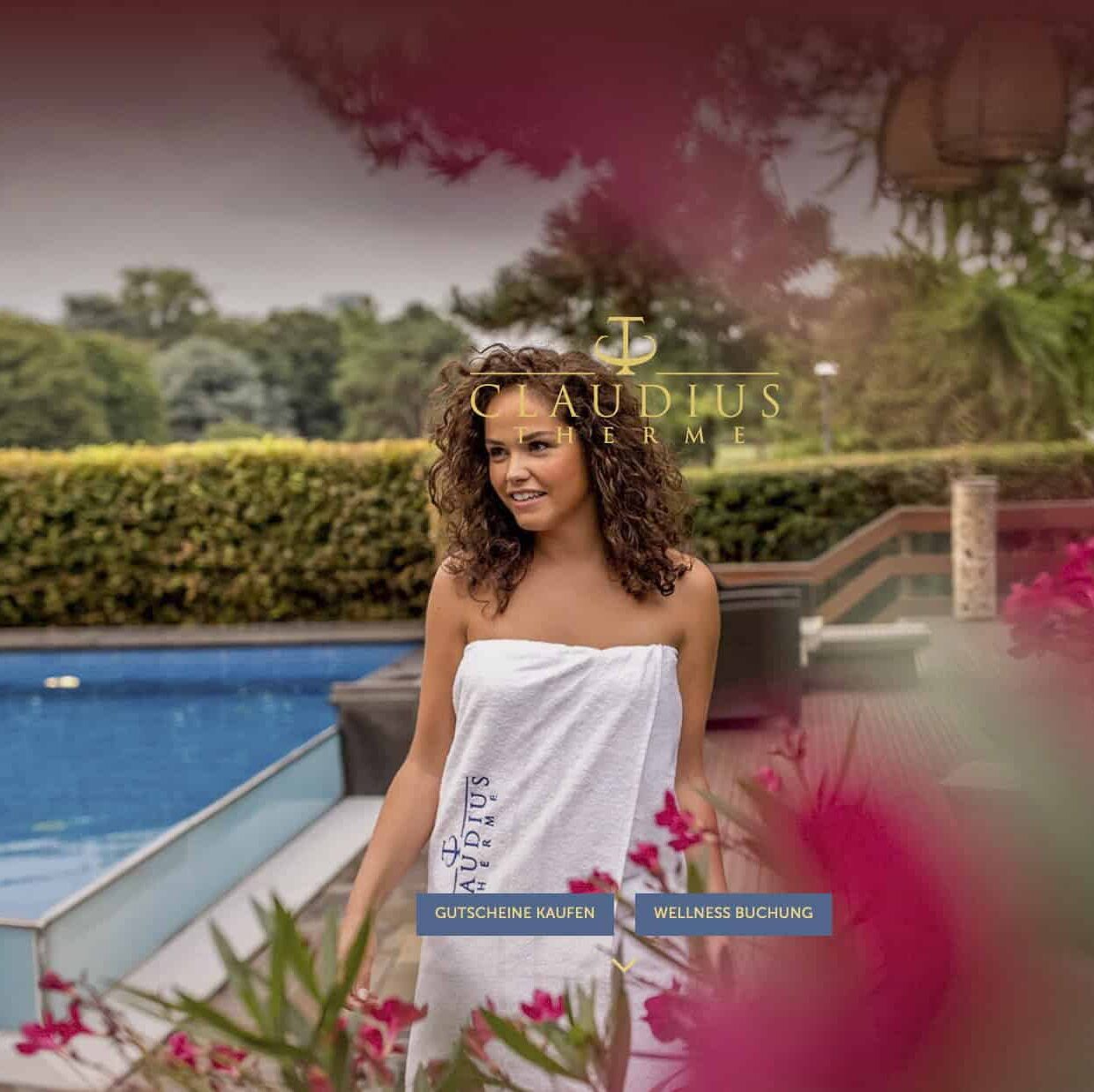 Eine lächelnde, in ein weißes Handtuch gehüllte Frau steht an einem Pool mit grünen Bäumen im Hintergrund und rosa Blumen im Vordergrund. Über ihr erscheint das Logo "Claudius Therme". © Fotografie Tomas Rodriguez