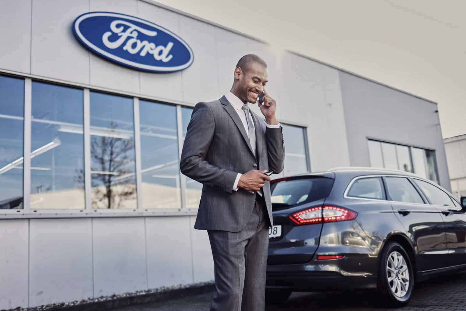 Ein Mann in einem grauen Anzug steht vor einem Ford-Händler, lächelt und telefoniert, während hinter ihm ein graues Auto parkt und das Ford-Logo auf dem Gebäude zu sehen ist. © Fotografie Tomas Rodriguez