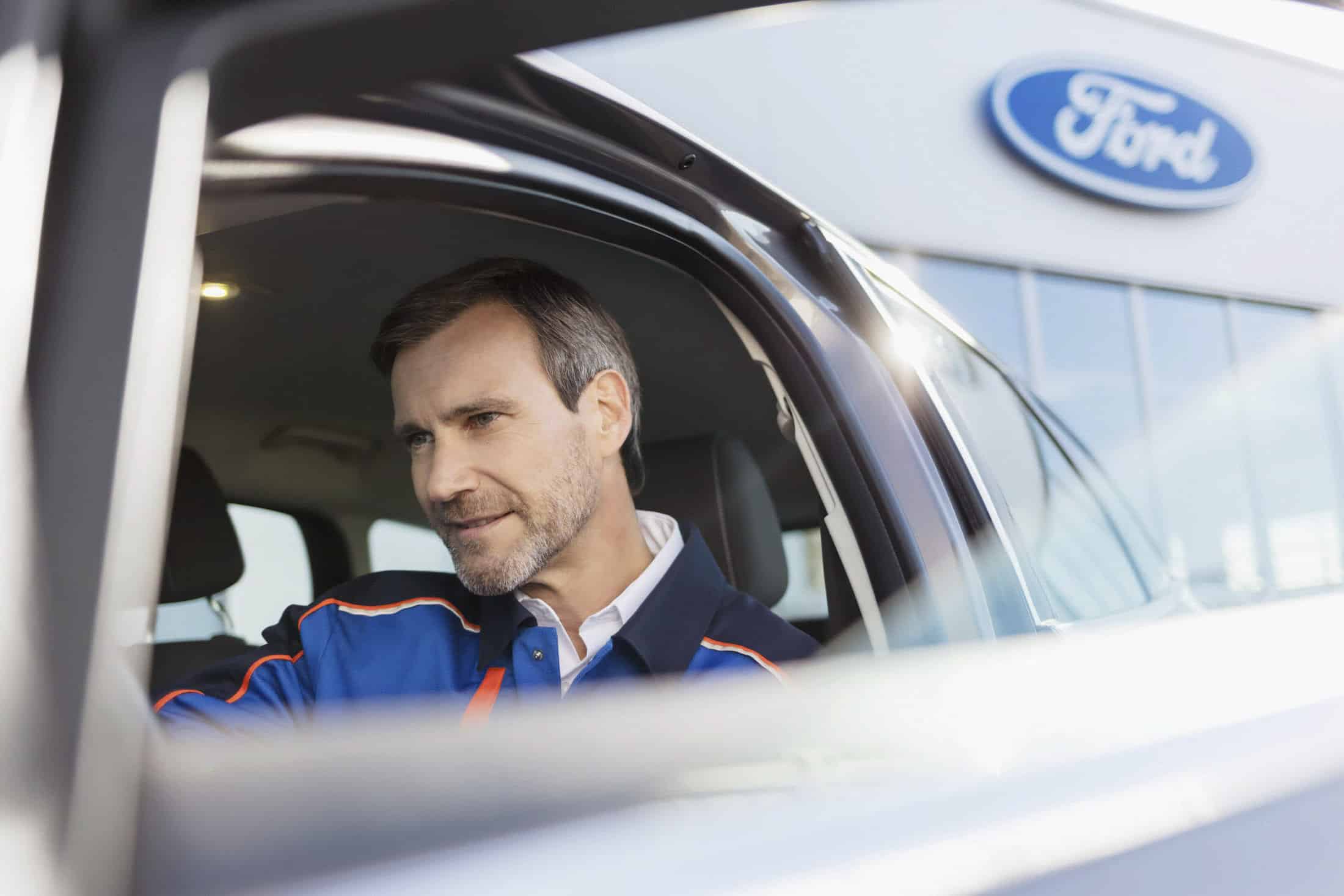 Ein Mann in einer blauen Jacke sitzt in einem Auto und schaut auf etwas, das vor ihm liegt. Das Foto wurde von außen aufgenommen, und auf einem Gebäude im Hintergrund ist ein Ford-Logo zu sehen. © Fotografie Tomas Rodriguez