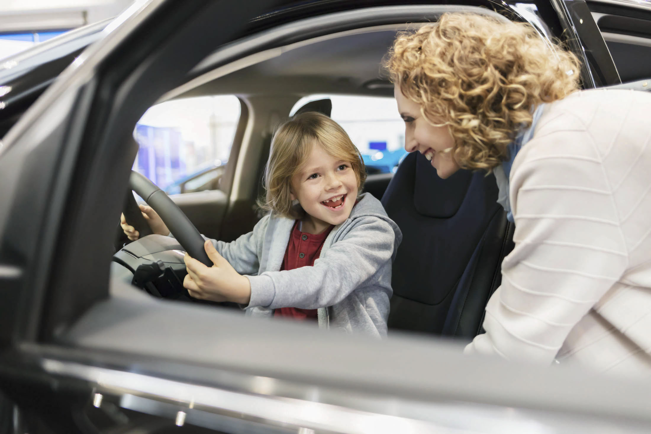 Ein lächelndes Kind sitzt auf dem Fahrersitz eines Autos und hält das Lenkrad fest, während sich eine Frau durch die geöffnete Tür hereinbeugt und das Kind anlächelt. © Fotografie Tomas Rodriguez