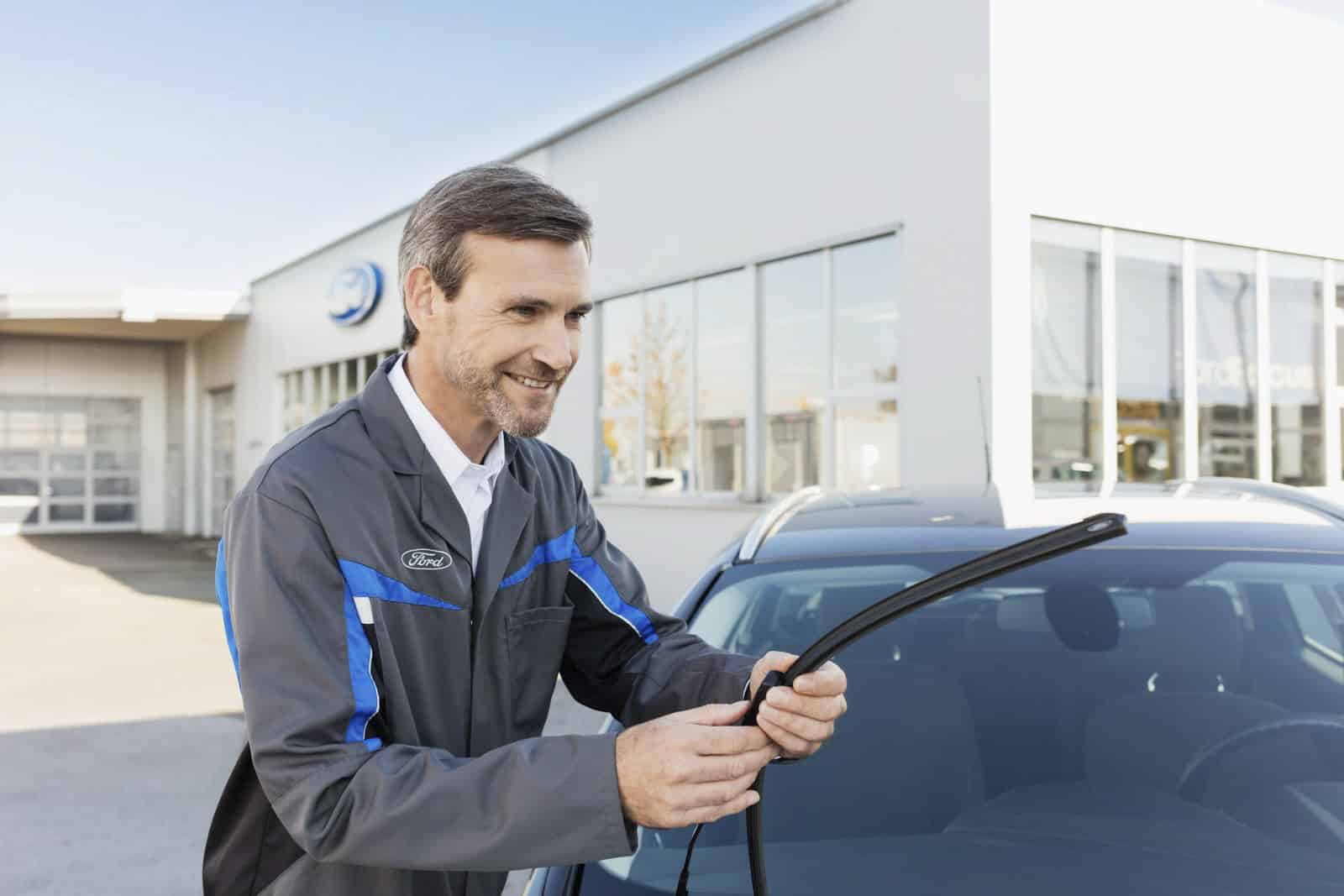 Ein lächelnder Automechaniker in einer grau-blauen Uniform montiert an einem sonnigen Tag ein Scheibenwischerblatt an einem Auto vor einem modernen Autohausgebäude. © Fotografie Tomas Rodriguez