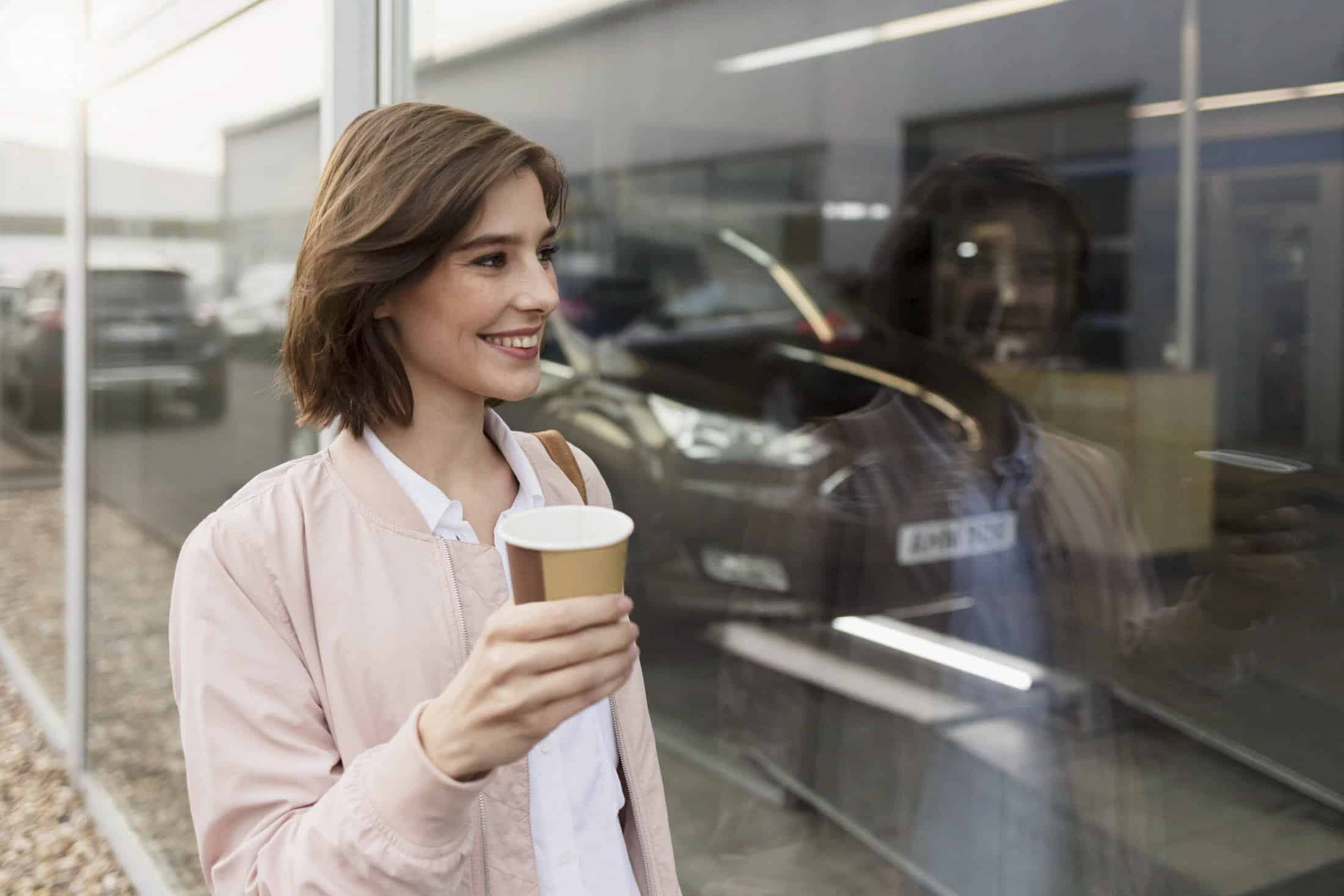 Eine Frau mit kurzen braunen Haaren, die eine hellrosa Jacke trägt, lächelt und hält einen Kaffeebecher aus Papier. Sie steht neben einem Glasfenster, in dem sich ihr Spiegelbild spiegelt. © Fotografie Tomas Rodriguez