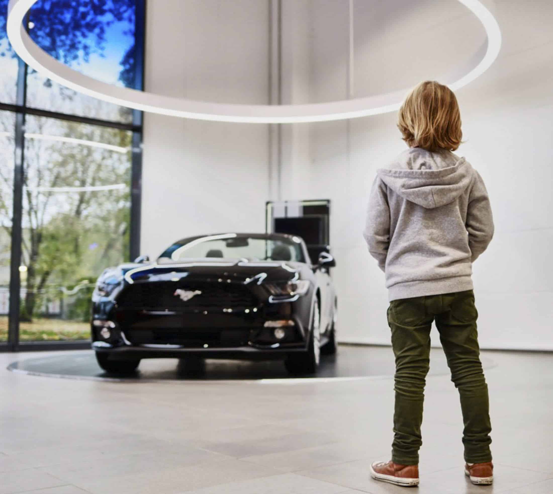 Ein Kind in einem grauen Kapuzenpulli und einer grünen Hose steht in einem modernen Ausstellungsraum vor einem schwarzen Ford Mustang, der unter einer runden Deckenleuchte ausgestellt ist und dessen große Fenster Bäume zeigen. © Fotografie Tomas Rodriguez