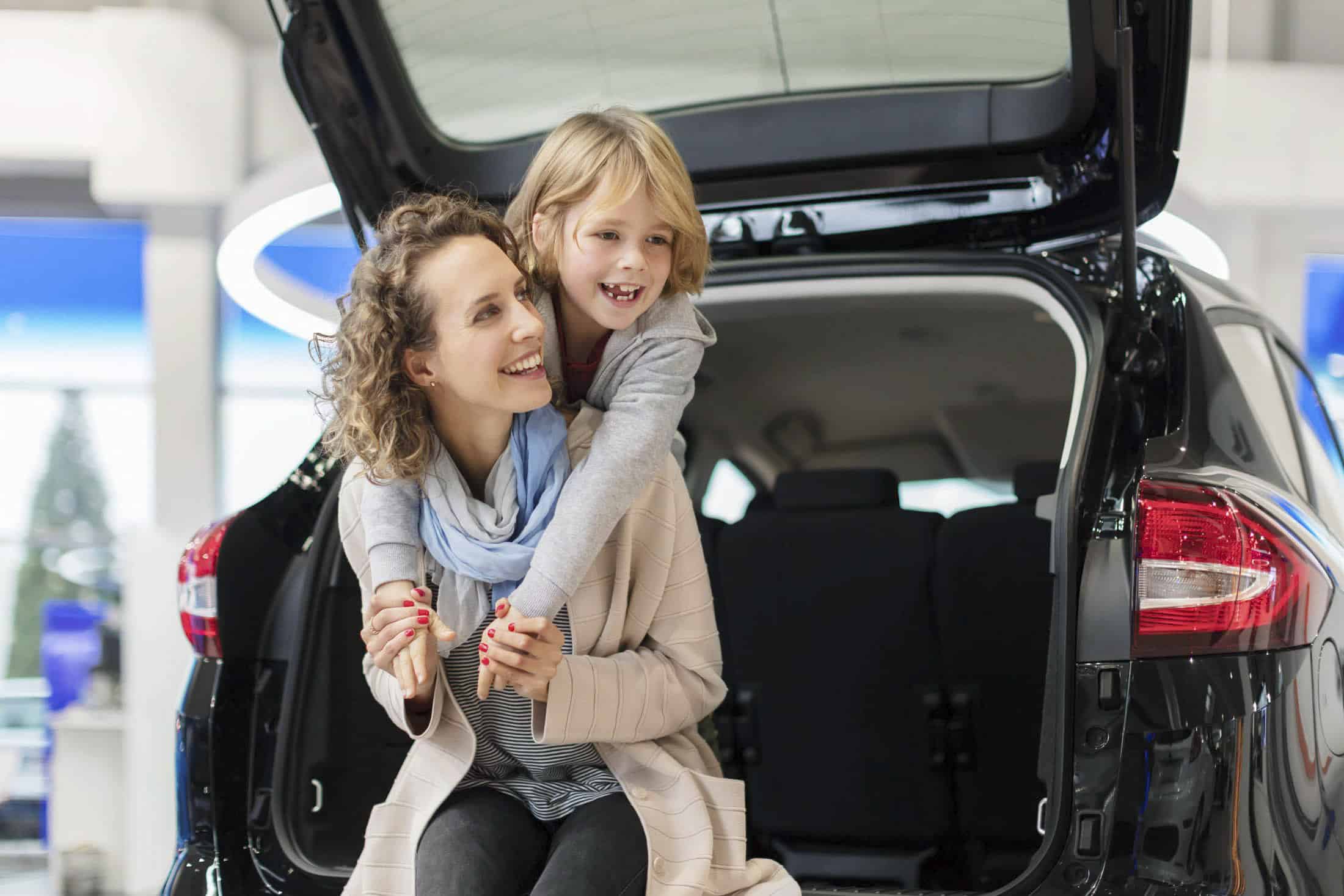Eine lächelnde Frau sitzt im offenen Kofferraum eines Autos, während ein glückliches Kind sie von hinten umarmt. Beide sehen fröhlich aus und sind leger gekleidet. Das Auto ist in einem Innenraum geparkt, was auf einen Ausstellungsraum oder ein Autohaus hindeutet. © Fotografie Tomas Rodriguez