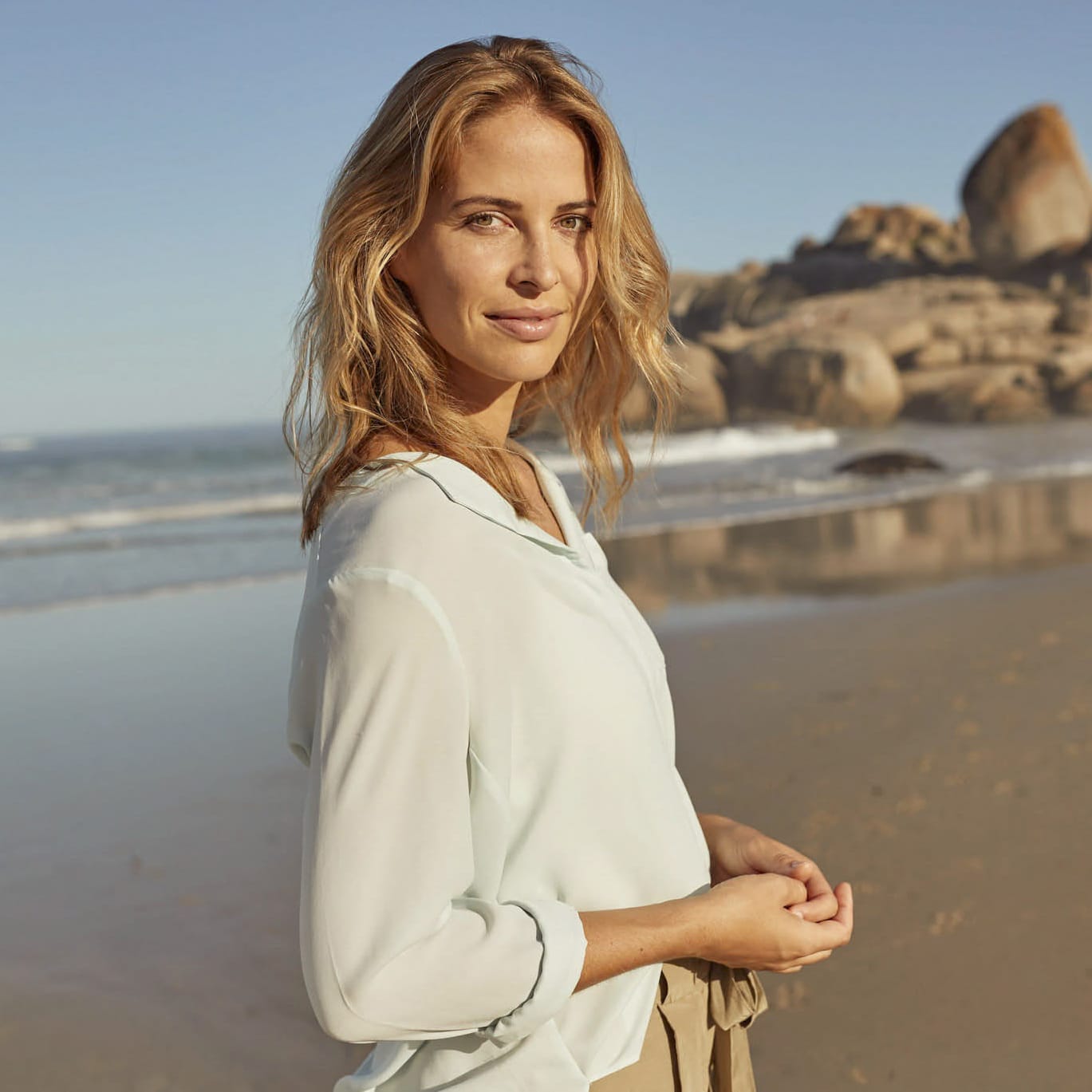 Eine Frau mit blondem Haar und heller Bluse steht an einem Sandstrand in der Nähe des Meeres, mit Felsen im Hintergrund, und blickt ruhig in die Kamera unter einem klaren blauen Himmel. © Fotografie Tomas Rodriguez