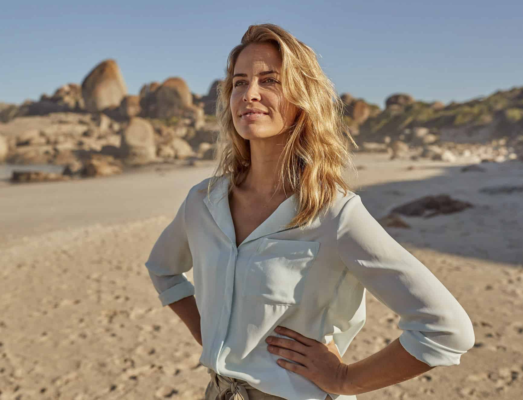 Eine Frau mit blondem Haar steht an einem Sandstrand mit großen Felsen im Hintergrund, trägt eine hellblaue Bluse und blickt selbstbewusst in die Ferne unter einem klaren Himmel. © Fotografie Tomas Rodriguez