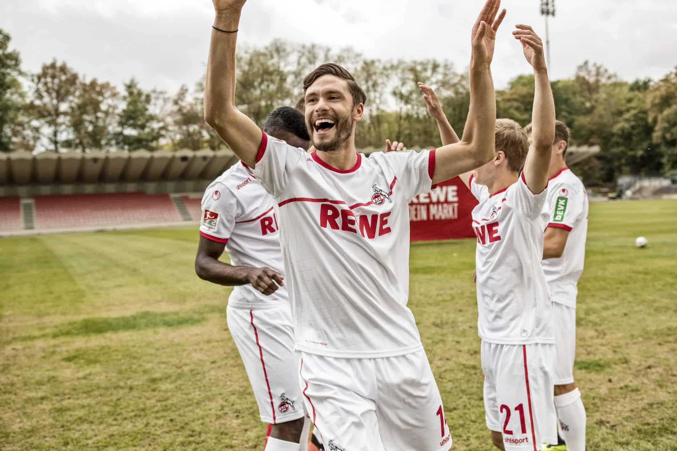 Eine Gruppe männlicher Fußballspieler in weißen Trikots feiert auf einem Feld, lächelt und hebt die Arme. Im Hintergrund sind die Sitzplätze und Bäume des Stadions zu sehen. © Fotografie Tomas Rodriguez