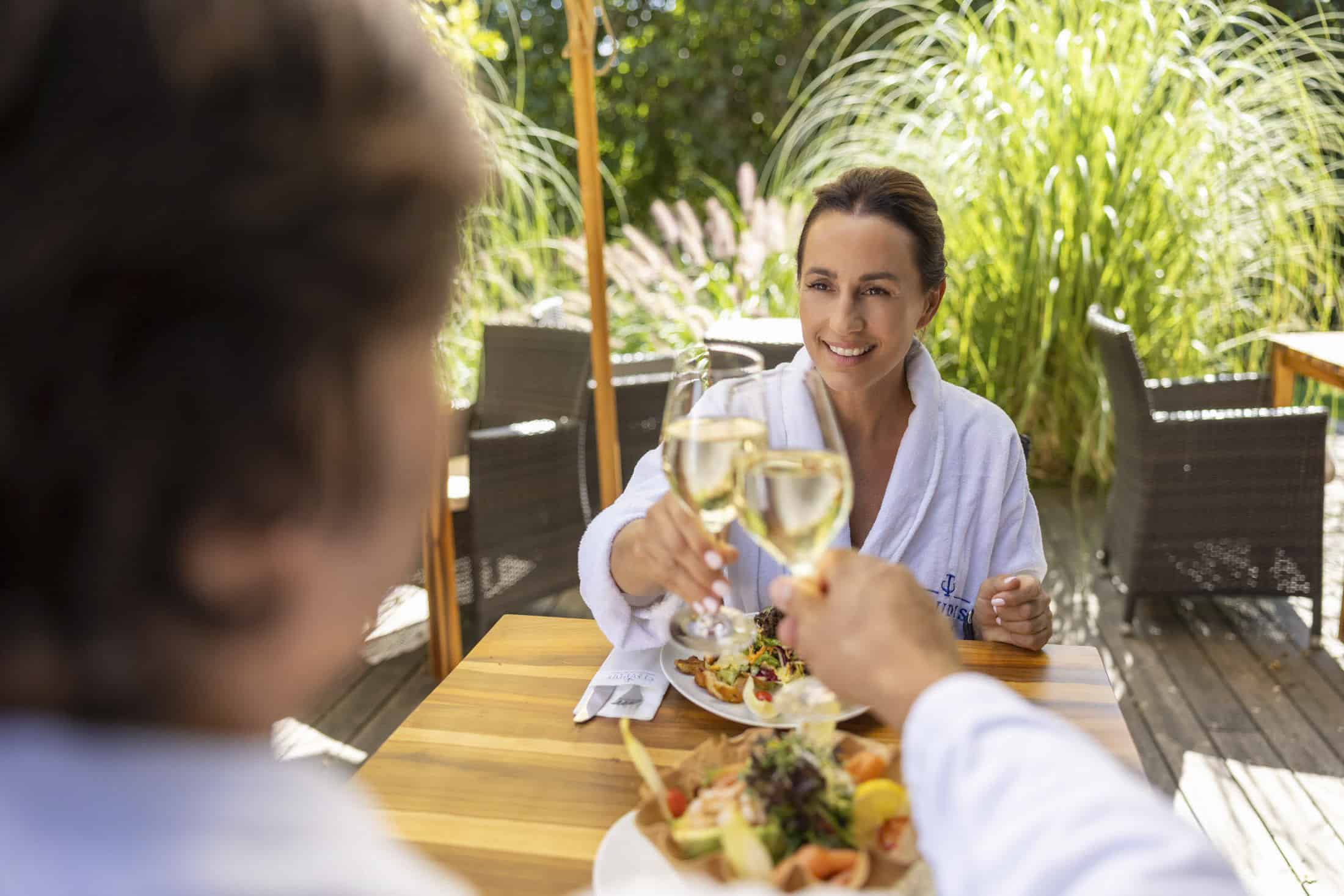 Zwei Personen in weißen Gewändern sitzen an einem Holztisch im Freien, lächeln und stoßen mit einem Glas Weißwein an. Vor ihnen stehen Teller mit frischem Salat und anderen Speisen, im Hintergrund ist üppiges Grün zu sehen. © Fotografie Tomas Rodriguez