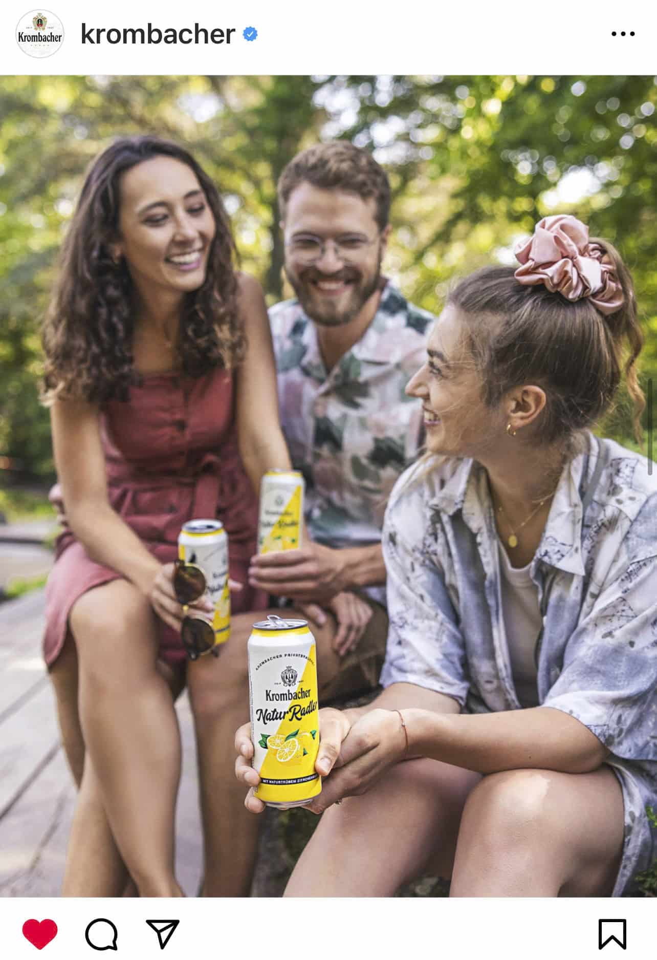 Drei lächelnde junge Erwachsene sitzen im Freien auf einer Bank, halten Dosen von Krombacher Lemon Radler und genießen einen sonnigen Tag. Im unscharfen Hintergrund sind Bäume und Grünflächen zu sehen. © Fotografie Tomas Rodriguez