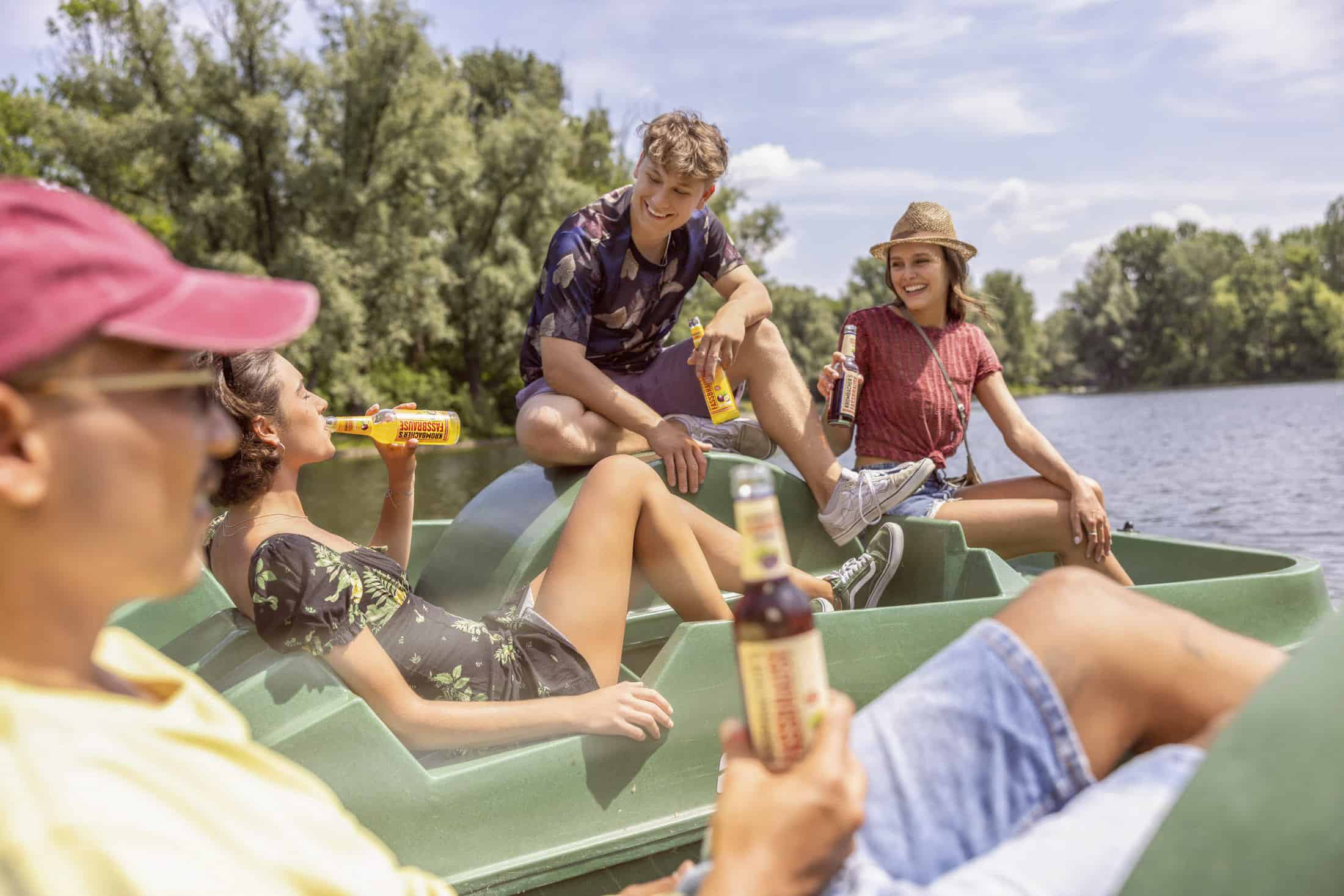 Vier junge Erwachsene entspannen sich auf einem Tretboot auf einem See und genießen Getränke in Flaschen an einem sonnigen Tag. Im Hintergrund sind Bäume und blauer Himmel zu sehen, die eine fröhliche, sommerliche Atmosphäre schaffen. © Fotografie Tomas Rodriguez