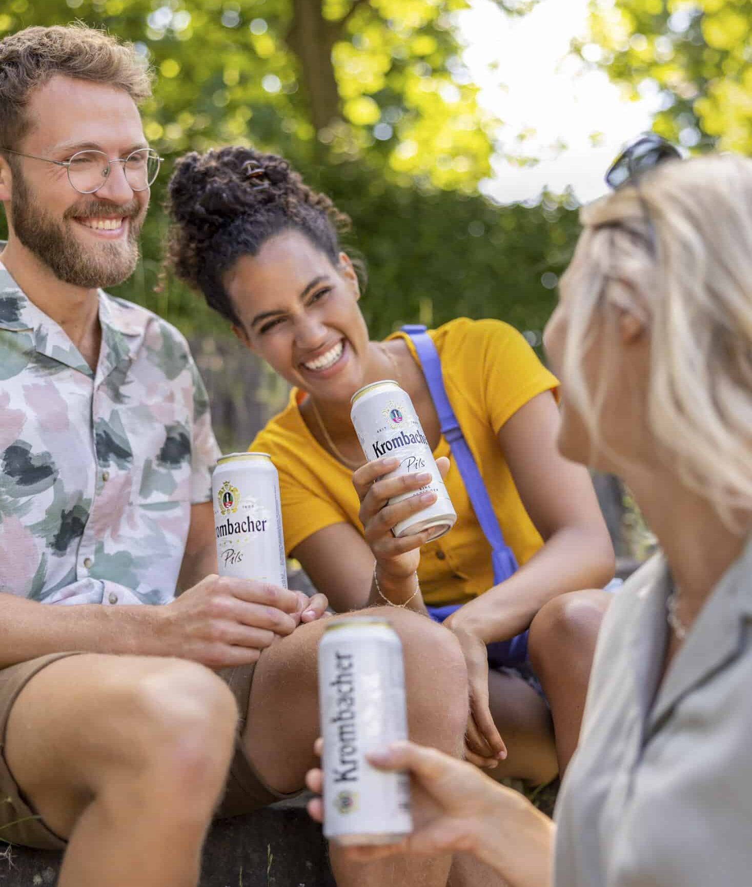 Drei Freunde sitzen an einem sonnigen Tag im Freien, lächeln und halten Dosen von Krombacher Bier in der Hand. Im Hintergrund sind Bäume und Grünflächen zu sehen, und die Gruppe scheint einen entspannten Moment miteinander zu genießen. © Fotografie Tomas Rodriguez