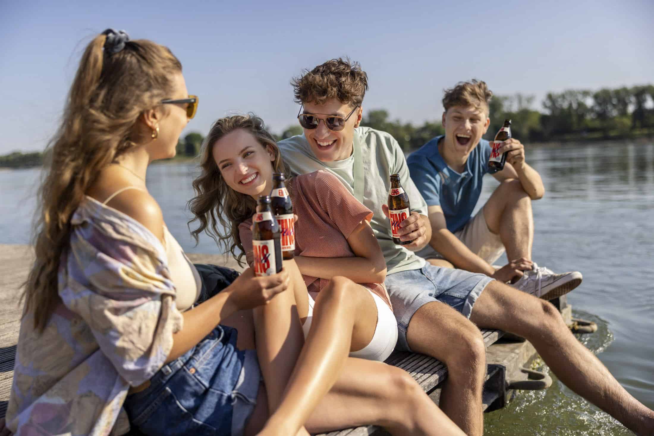 Vier junge Erwachsene sitzen auf einem hölzernen Steg an einem See, lächeln und lachen und halten Bierflaschen in der Hand. Es ist ein sonniger Tag und sie wirken entspannt und glücklich und genießen die Gesellschaft des anderen am Wasser. © Fotografie Tomas Rodriguez