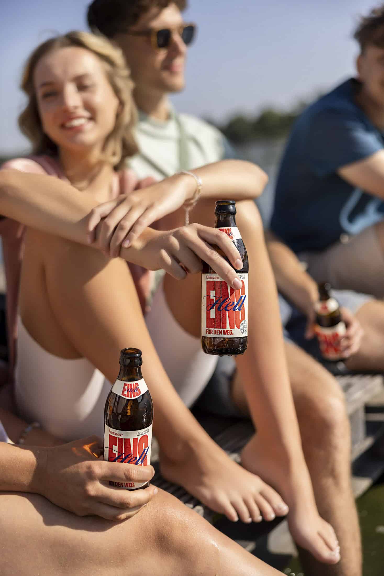 Drei junge Erwachsene sitzen im Freien im Sonnenlicht, lächeln und entspannen sich, jeder hält eine Flasche ENO Golden Weiss Bier. Der Hintergrund ist leicht verschwommen und suggeriert eine zwanglose, sommerliche Umgebung in der Nähe von Wasser oder Natur. © Fotografie Tomas Rodriguez