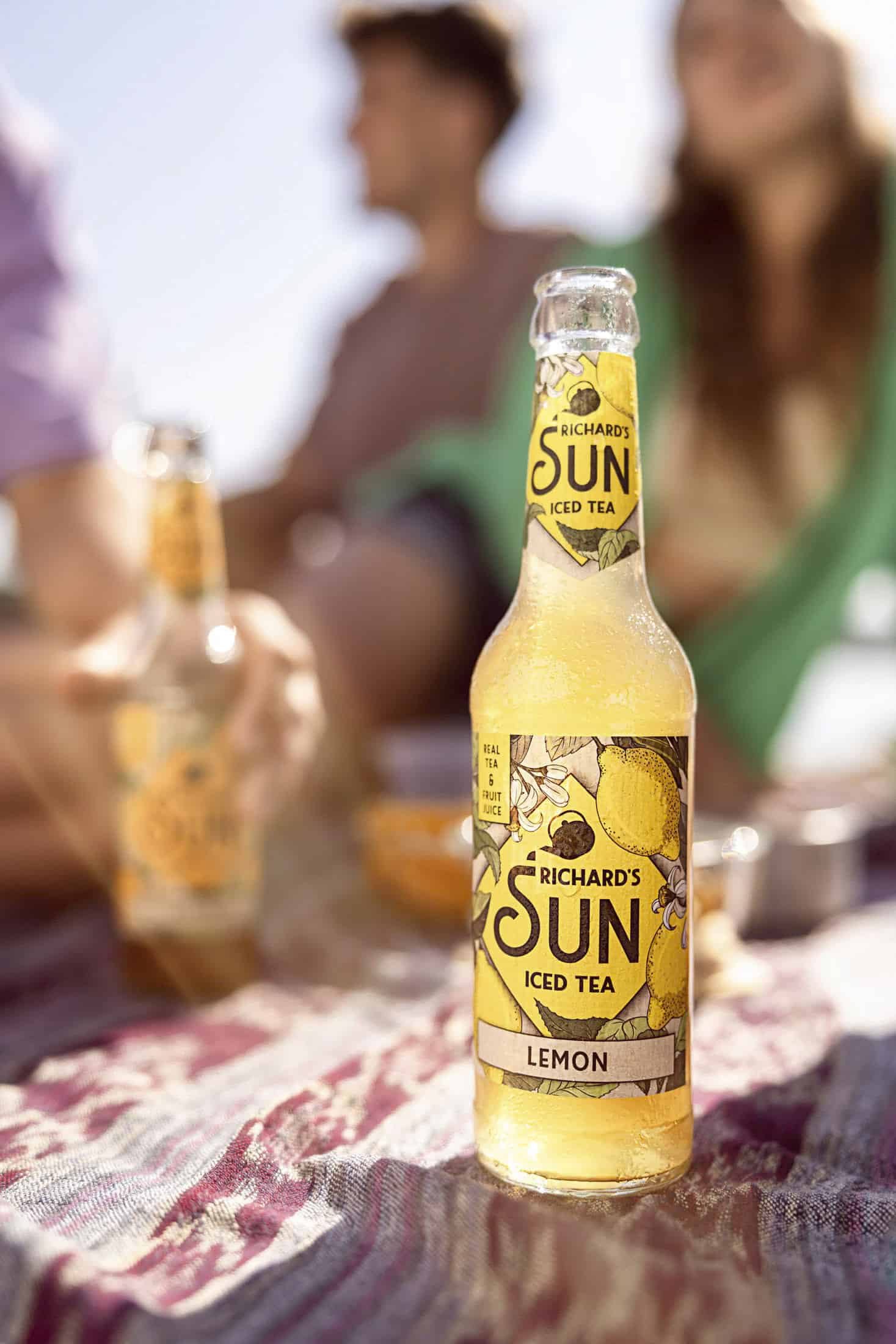 Eine Flasche Richard's Sun Lemon Iced Tea steht auf einer gemusterten Picknickdecke im Freien. Zwei Personen und eine weitere Flasche sind unscharf im Hintergrund zu erkennen, was auf ein entspanntes, sonniges Picknick hinweist. © Fotografie Tomas Rodriguez