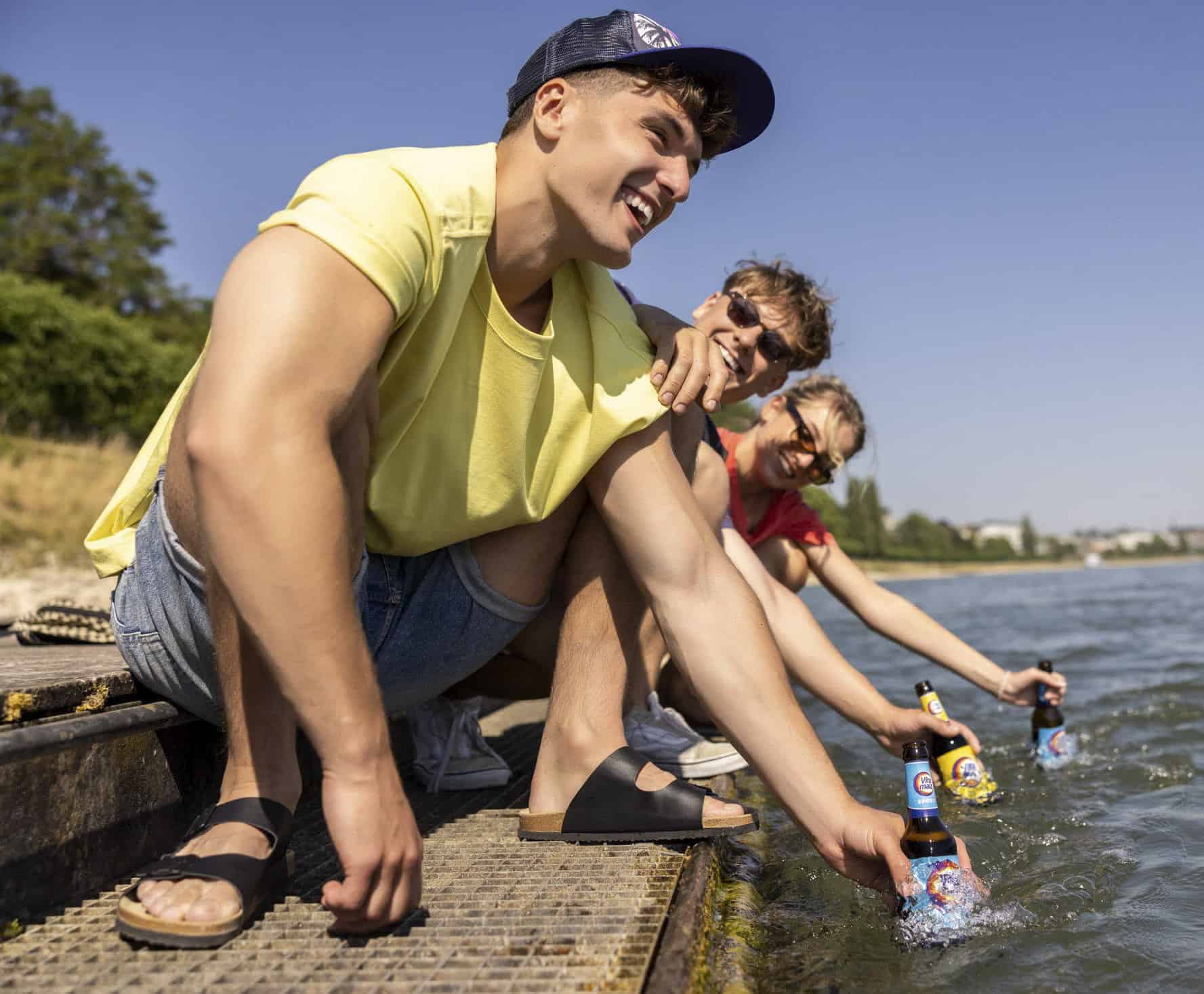 Drei junge Erwachsene sitzen an einem sonnigen Tag auf einem Steg am Wasser, lächeln und kühlen ihre Bierflaschen im See. Sie tragen legere Sommerkleidung und Sonnenbrillen und genießen gemeinsam einen entspannten Moment im Freien. © Fotografie Tomas Rodriguez