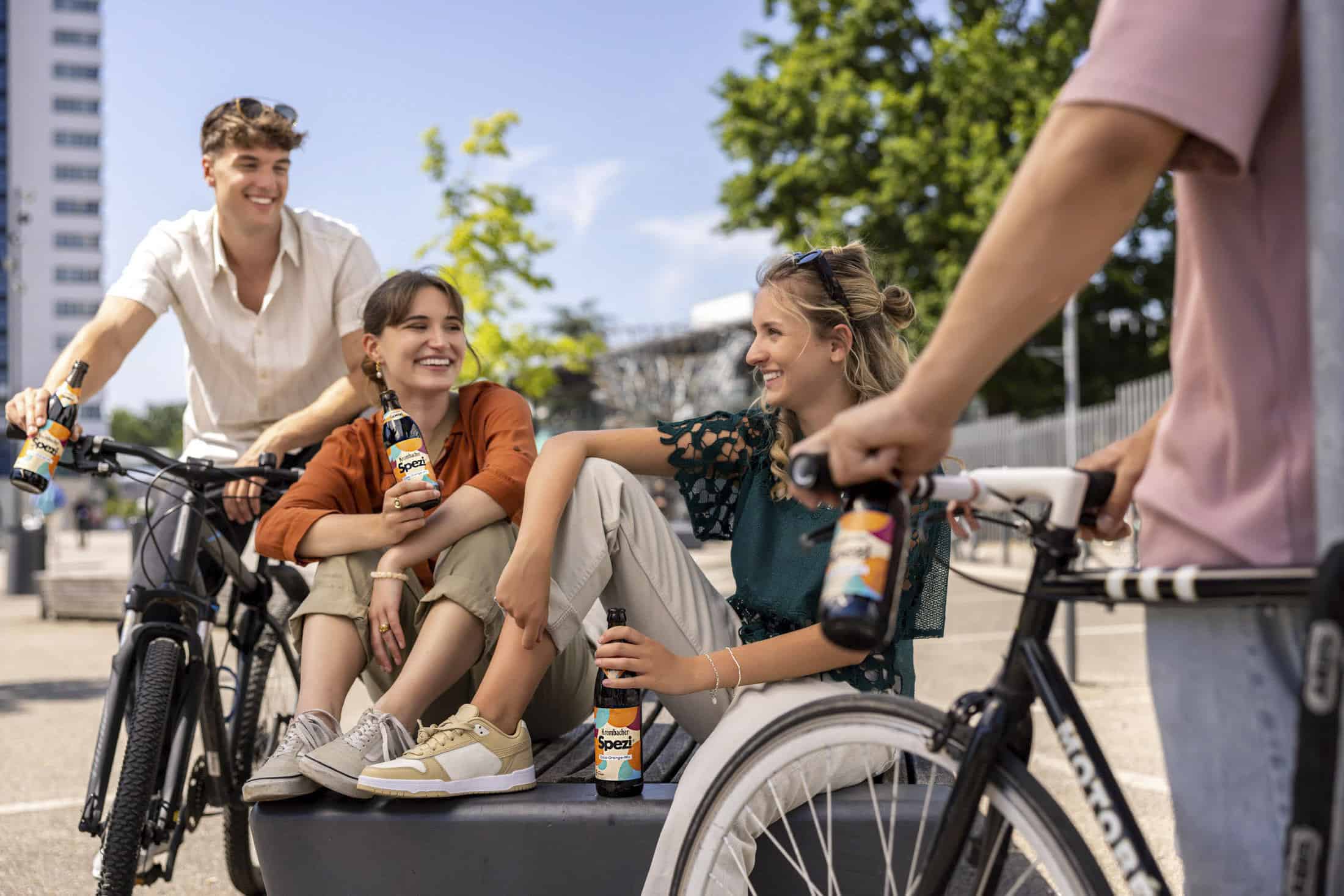 Vier junge Erwachsene mit Fahrrädern unterhalten sich an einem sonnigen Tag im Freien, lächeln und halten Flaschen mit Spezi-Soda in der Hand. Zwei sitzen auf einer Bank, während zwei in der Nähe mit ihren Fahrrädern stehen. Im Hintergrund sind Bäume und ein hohes Gebäude zu sehen. © Fotografie Tomas Rodriguez