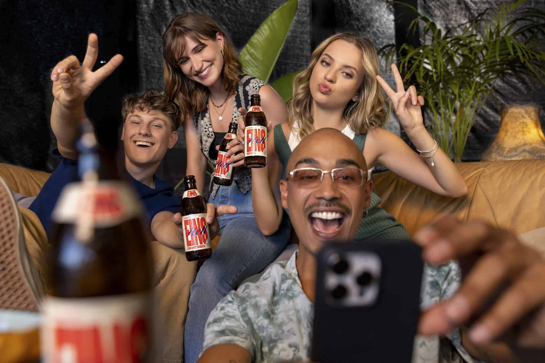 Vier Freunde sitzen auf einer Couch, lächeln und machen Friedenszeichen, während sie ein Selfie machen. Jeder von ihnen hält eine Flasche Limonade in der Hand, und im Hintergrund sind Pflanzen zu sehen, die eine entspannte und fröhliche Atmosphäre schaffen. © Fotografie Tomas Rodriguez