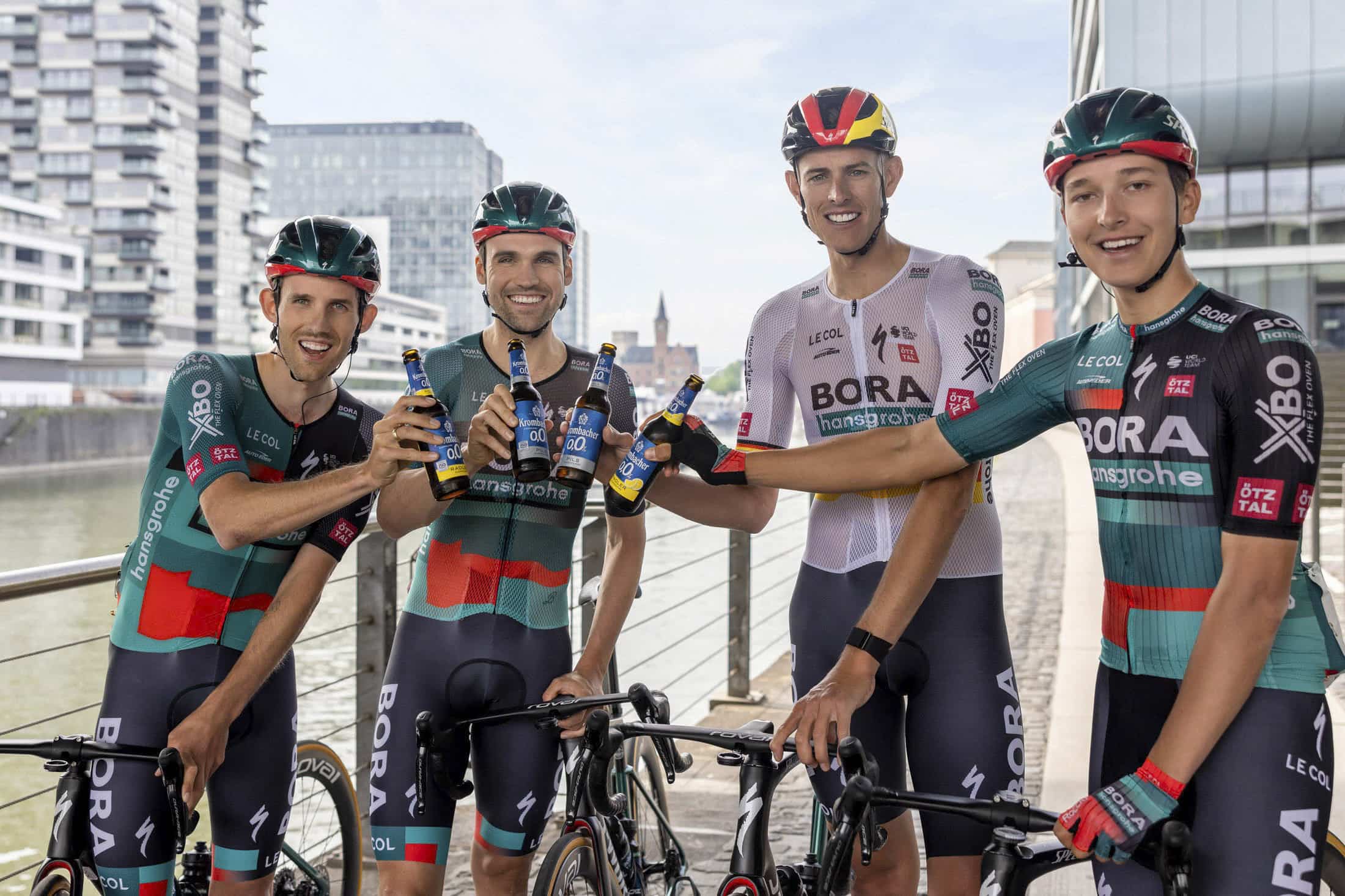 Vier Radfahrer in BORA-hansgrohe Uniformen lächeln und stoßen mit Flaschen an, während sie mit ihren Fahrrädern auf einer Uferpromenade in einem städtischen Gebiet stehen. © Fotografie Tomas Rodriguez