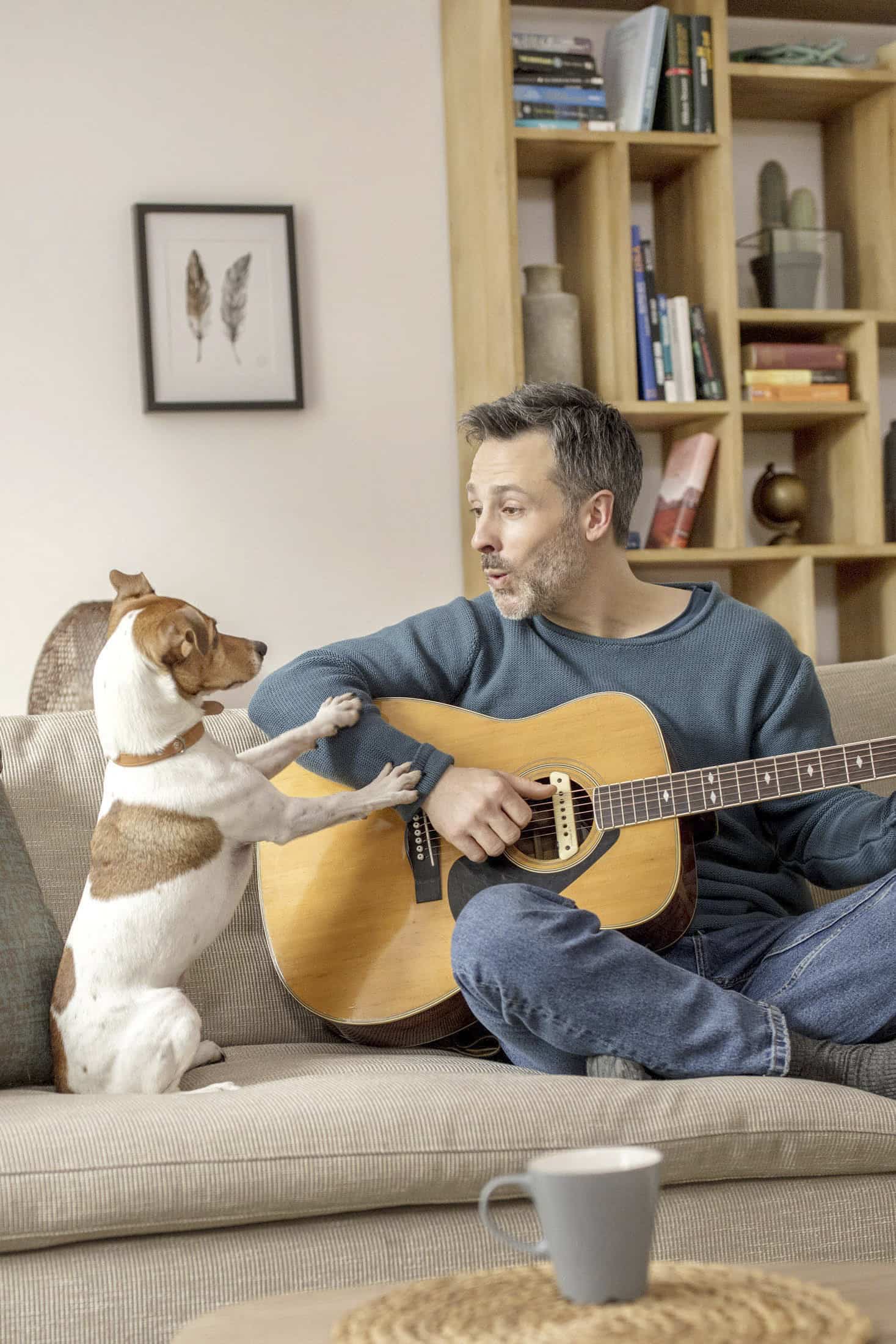 Ein Mann sitzt auf einer Couch und spielt Gitarre, während ein kleiner Hund mit seinen Vorderpfoten auf seinem Schoß ihn anschaut. In dem gemütlichen Wohnzimmer sind Regale, gerahmte Kunstwerke und ein Couchtisch mit einem Becher zu sehen. © Fotografie Tomas Rodriguez