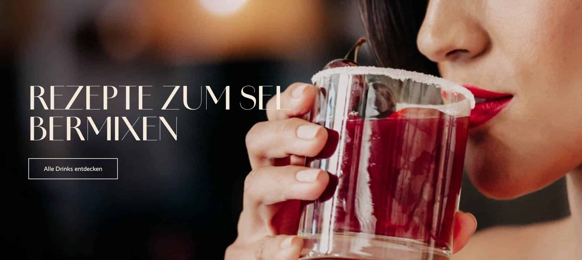 Eine Frau mit rotem Lippenstift hält ein Glas mit einem dunkelroten Cocktail mit Kirsch- und Zuckerrand, um einen Schluck zu nehmen. Auf dem Text-Overlay steht "Rezepte zum Selbermixen" und auf einer Schaltfläche steht "Alle Drinks entdecken". © Fotografie Tomas Rodriguez