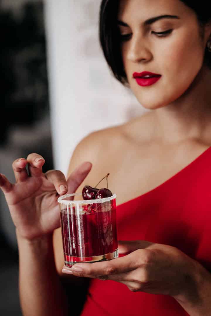 Eine Frau in einem roten Kleid hält ein Glas mit einem roten Cocktail, garniert mit Kirschen. Sie schaut auf das Getränk und berührt sanft das Glas. Sie hat dazu passenden roten Lippenstift und kurze dunkle Haare. © Fotografie Tomas Rodriguez