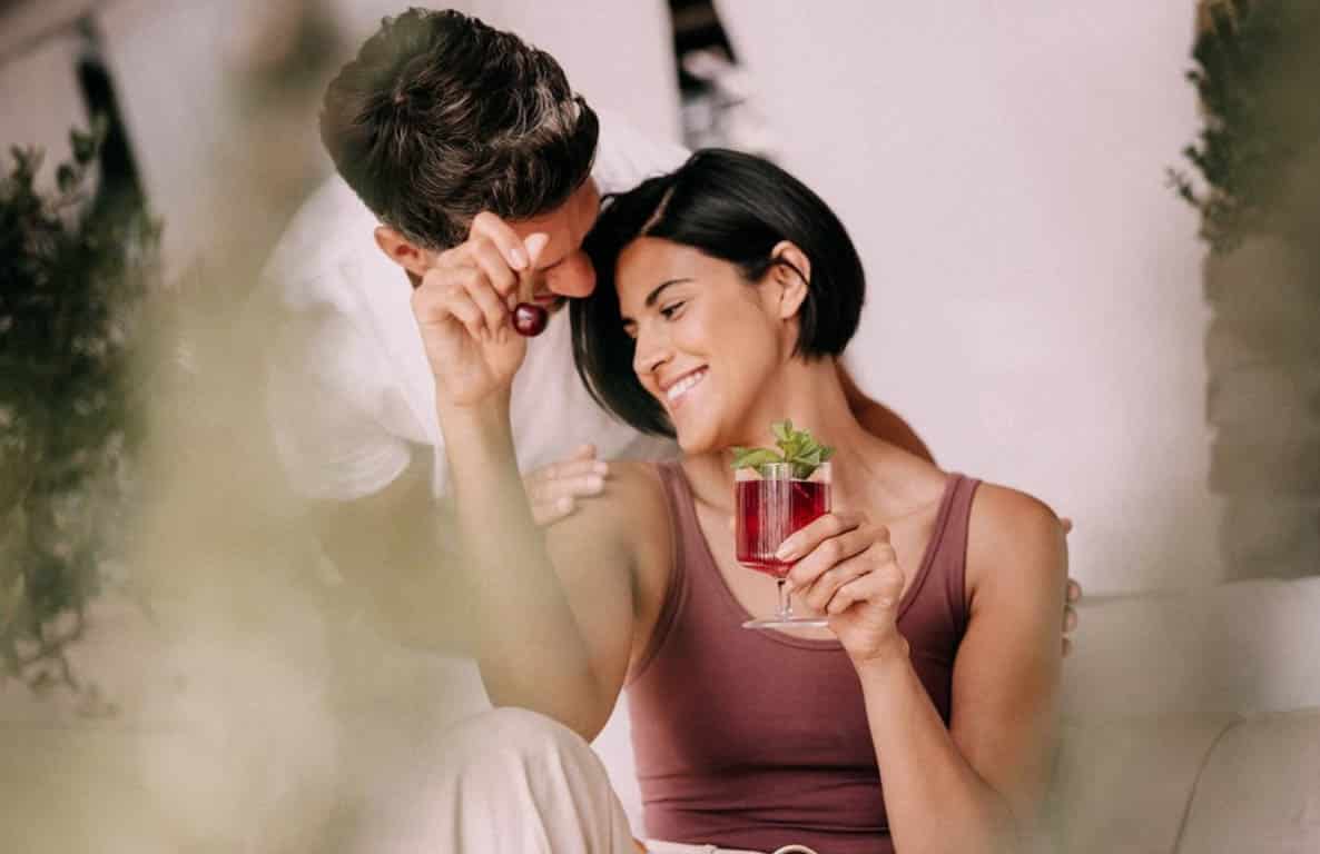 Ein lächelndes Paar sitzt eng beieinander. Die Frau hält ein Glas mit einem roten, mit Minze garnierten Getränk in der Hand, während der Mann sich an sie lehnt und eine Kirsche in der Hand hält. Sie wirken glücklich und entspannt in einer hellen, gemütlichen Umgebung. © Fotografie Tomas Rodriguez