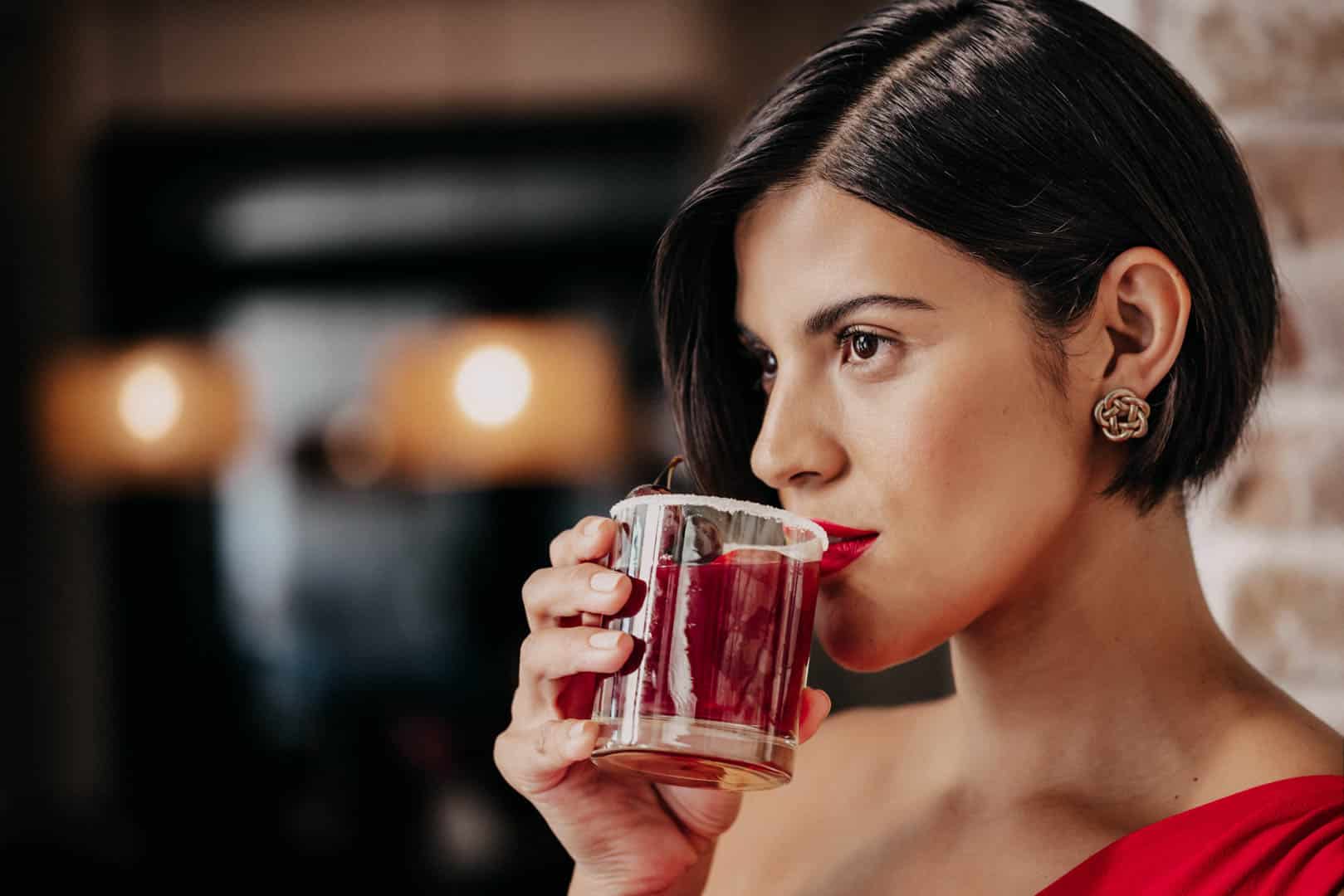 Eine Frau mit kurzen dunklen Haaren und rotem Lippenstift, die ein rotes Kleid und goldene Ohrringe trägt, hält ein rotes Getränk in der Hand und nippt daran in einem schwach beleuchteten Innenraum. © Fotografie Tomas Rodriguez