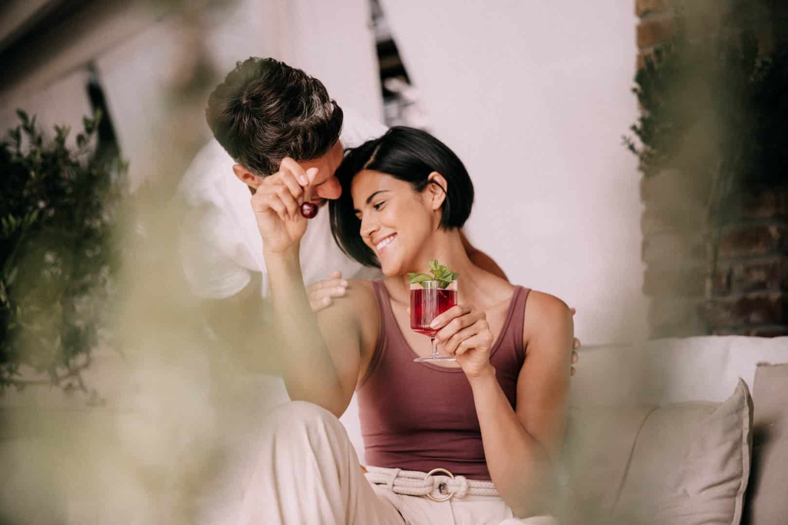 Eine lächelnde Frau, die ein rotes Getränk mit Minze in der Hand hält, sitzt auf einer Couch, während sich ein Mann dicht an sie heranlehnt und ihr spielerisch eine Kirsche ins Gesicht hält. Sie wirken glücklich und entspannt, umgeben von sanftem Grün. © Fotografie Tomas Rodriguez