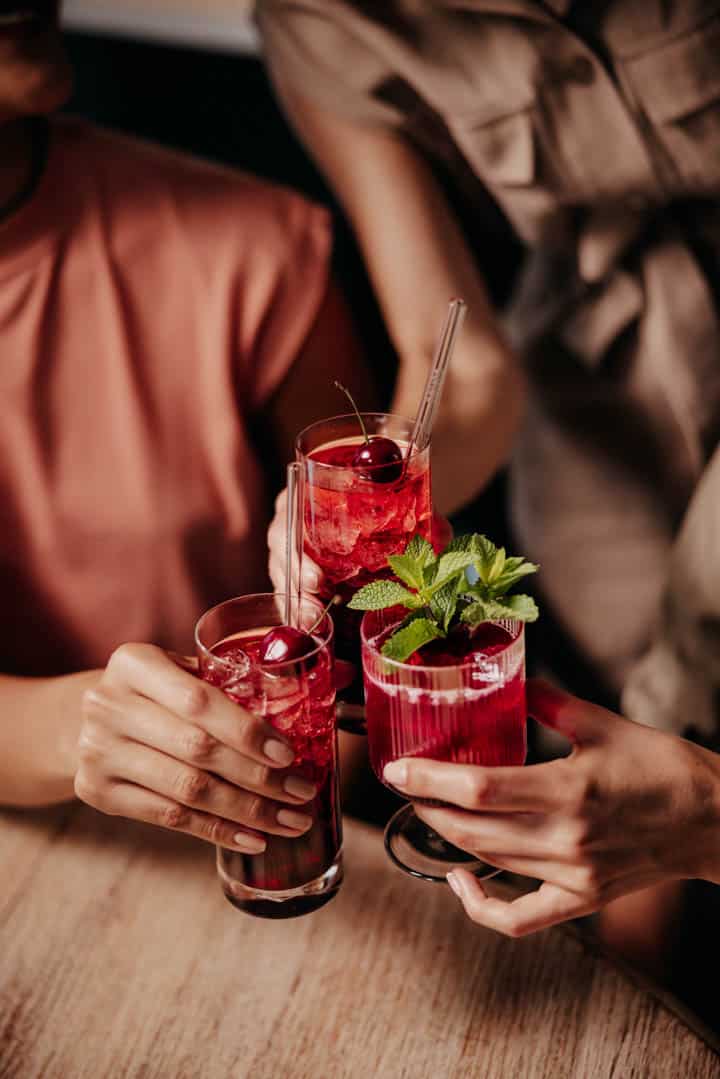 Drei Personen erheben Gläser mit leuchtend roten Cocktails, garniert mit Kirschen und Minze, und feiern gemeinsam an einem Holztisch. Die Szene ist warm beleuchtet und hebt ihre Hände und Getränke hervor. © Fotografie Tomas Rodriguez