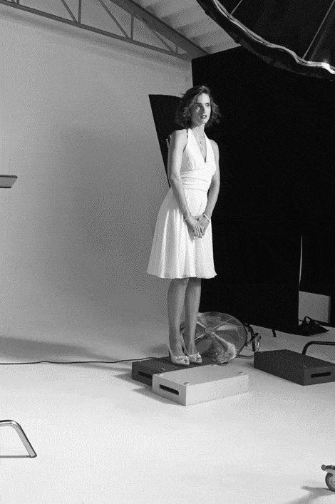 Eine Frau in einem weißen Kleid steht auf einem Podest in einem Fotostudio, umgeben von Beleuchtungsgeräten und einem Hintergrund. Sie blickt leicht zur Seite und hat die Hände vor sich verschränkt. © Fotografie Tomas Rodriguez