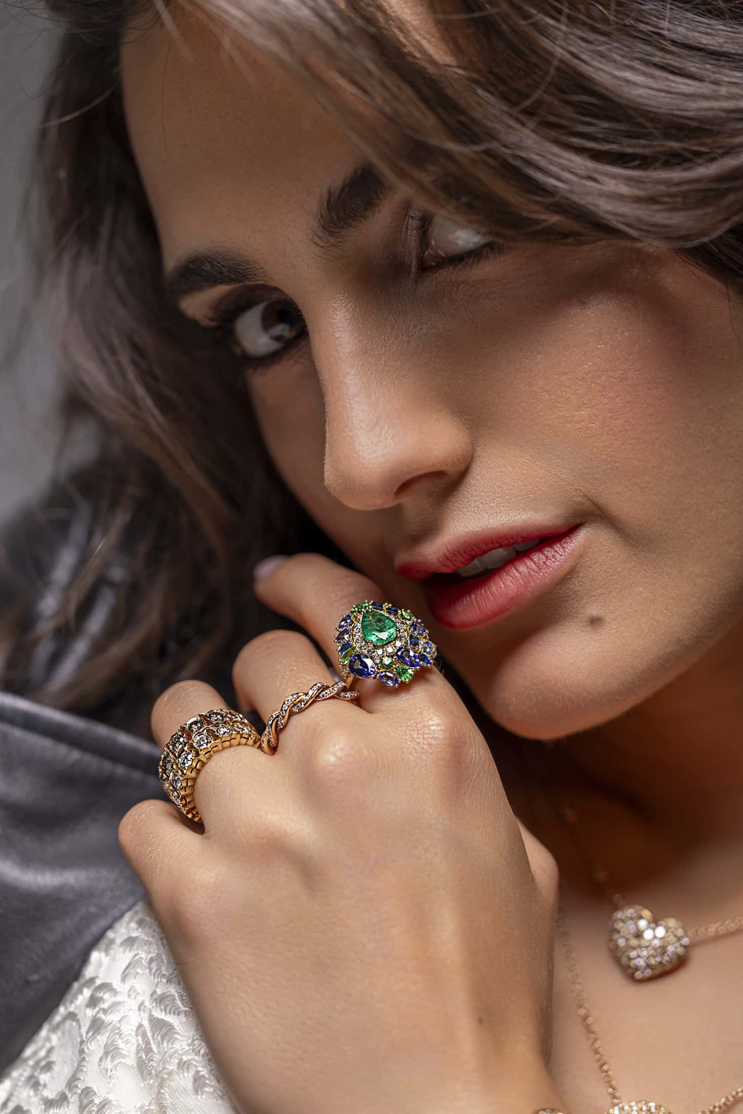 Eine Frau mit langen braunen Haaren blickt in die Kamera, die Hand vor dem Gesicht. Sie trägt auffällige Ringe mit bunten Edelsteinen und Gold, dazu eine Halskette und ein strukturiertes silbernes oder weißes Kleidungsstück. © Fotografie Tomas Rodriguez