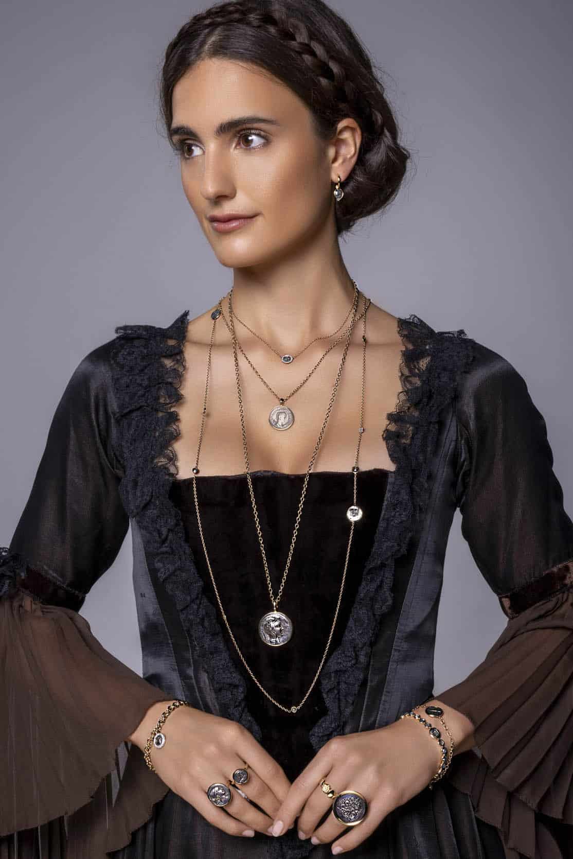 Eine Frau mit geflochtenen dunklen Haaren trägt ein schwarzes, gerüschtes Kleid und mehrlagige Halsketten mit Anhängern, dazu Ringe, Armbänder und Ohrringe. Sie blickt leicht zur Seite vor einem schlichten Hintergrund. © Fotografie Tomas Rodriguez