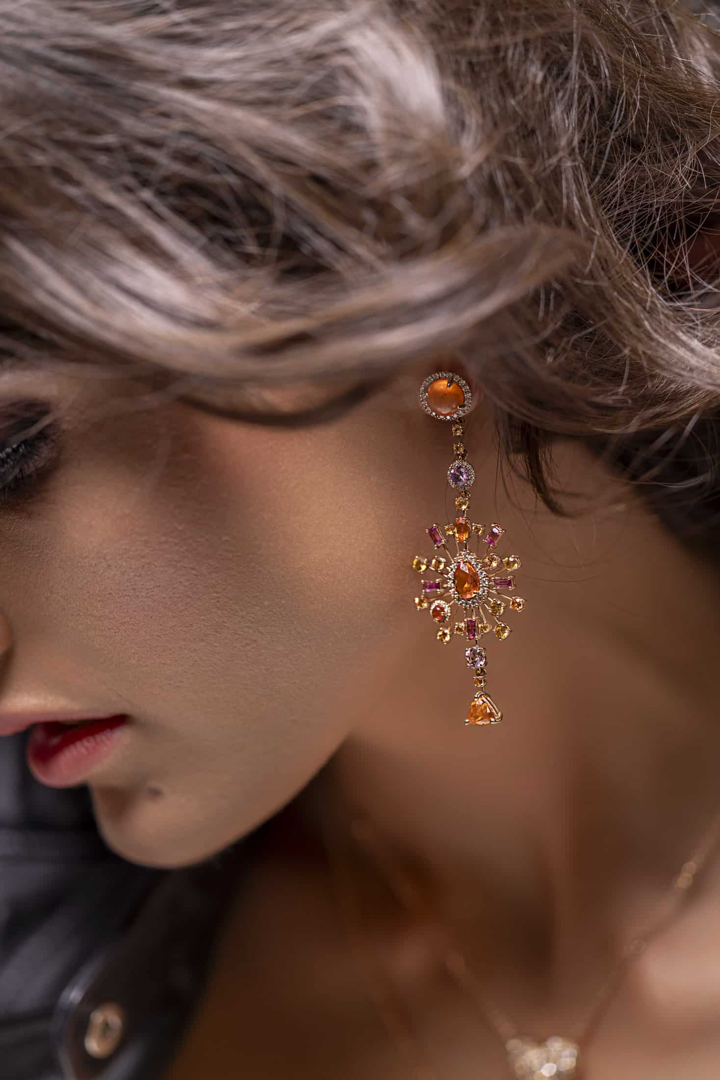 Eine Nahaufnahme des Gesichts einer Frau, wobei der Schwerpunkt auf ihrem gewellten braunen Haar, den roten Lippen, dem Schönheitsfleck in der Nähe ihres Mundes und einem detaillierten gold- und orangefarbenen, mit Juwelen besetzten Kronleuchter-Ohrring liegt. © Fotografie Tomas Rodriguez