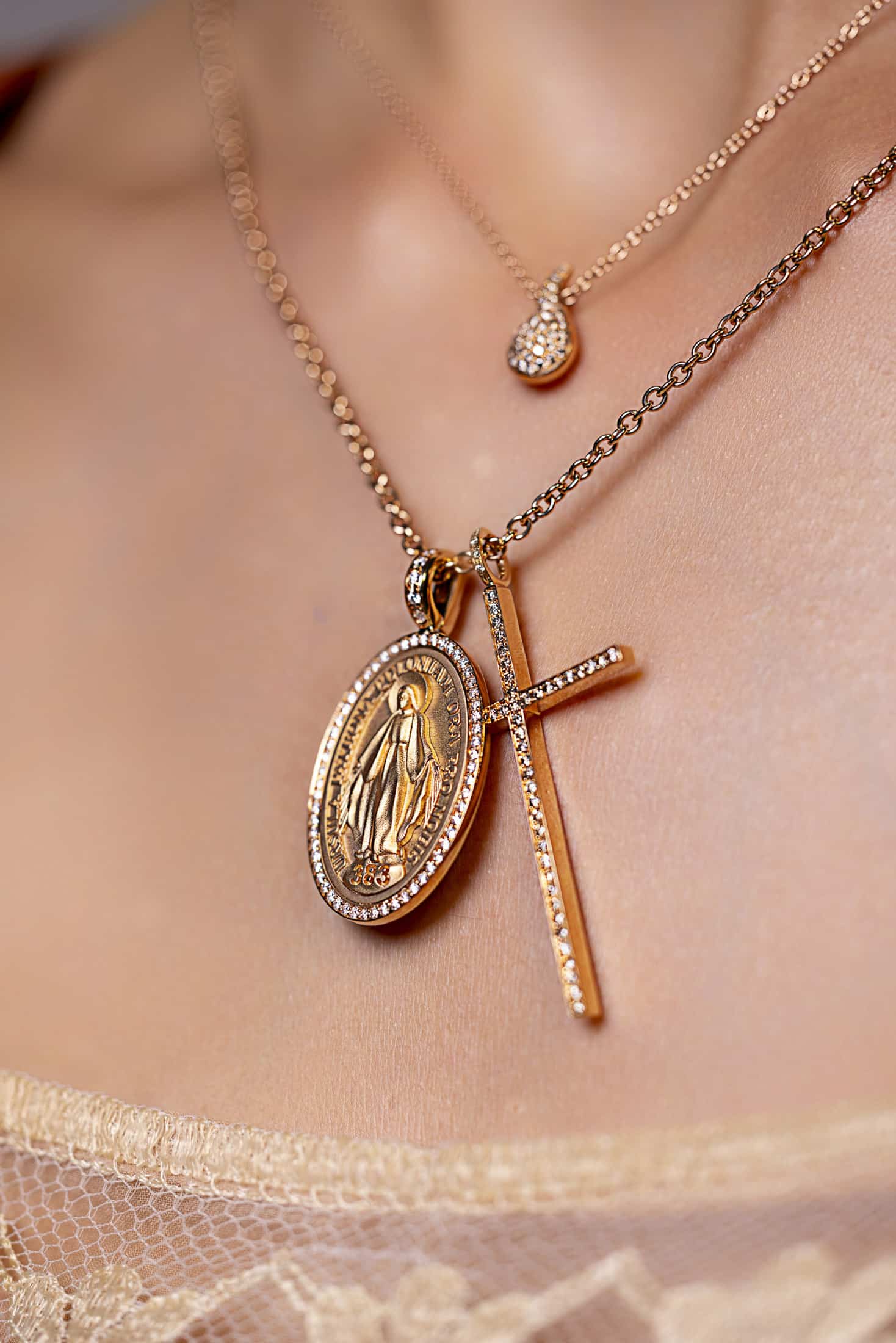 Nahaufnahme des Halses einer Person, die zwei goldene Halsketten trägt: eine mit einem ovalen religiösen Medaillon und einem Kreuzanhänger und eine andere mit einem kleinen, runden, funkelnden Anhänger. Die Person ist mit einem spitzenbesetzten Oberteil bekleidet. © Fotografie Tomas Rodriguez