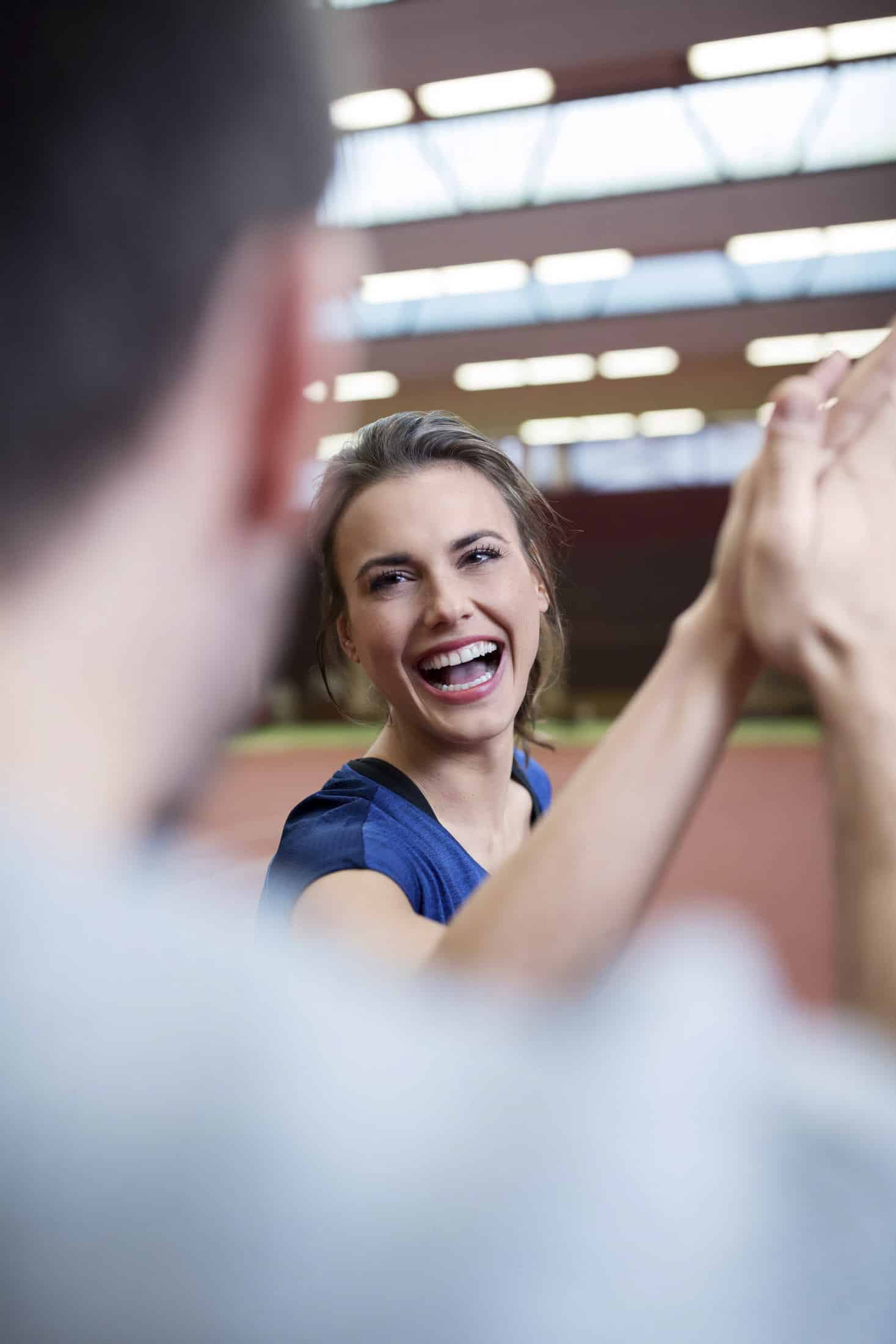 Eine Frau, die lächelt und einer unscharfen Person im Vordergrund ein "High Five" gibt, drinnen auf einem Sportplatz mit hellem Oberlicht. © Fotografie Tomas Rodriguez