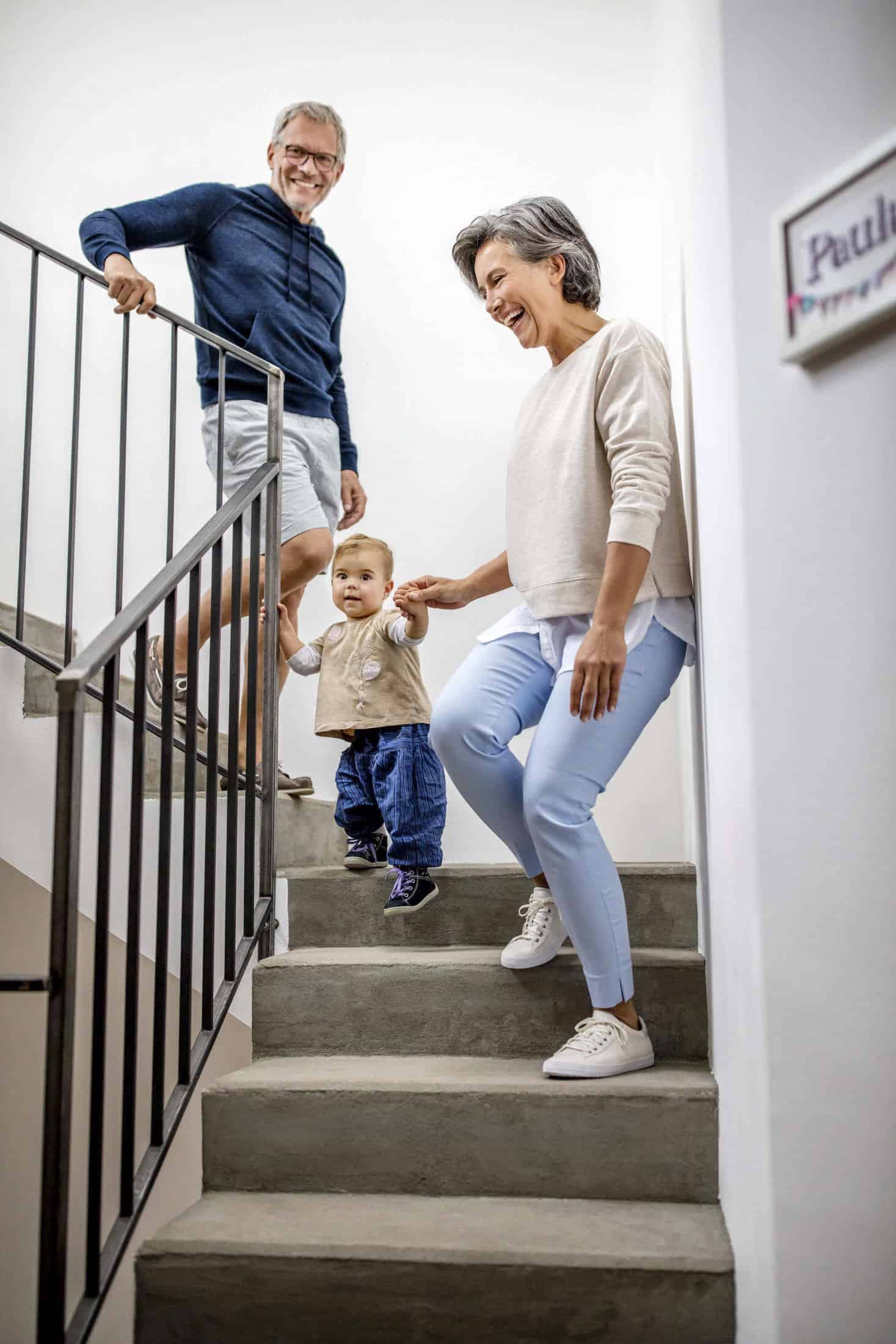Ein älteres Ehepaar hilft einem Kleinkind, eine Treppe hinunterzugehen. Die Frau hält das Kind an der Hand, während der Mann dicht hinterherläuft. Alle lächeln und scheinen glücklich in ihrem Haus zu sein. © Fotografie Tomas Rodriguez