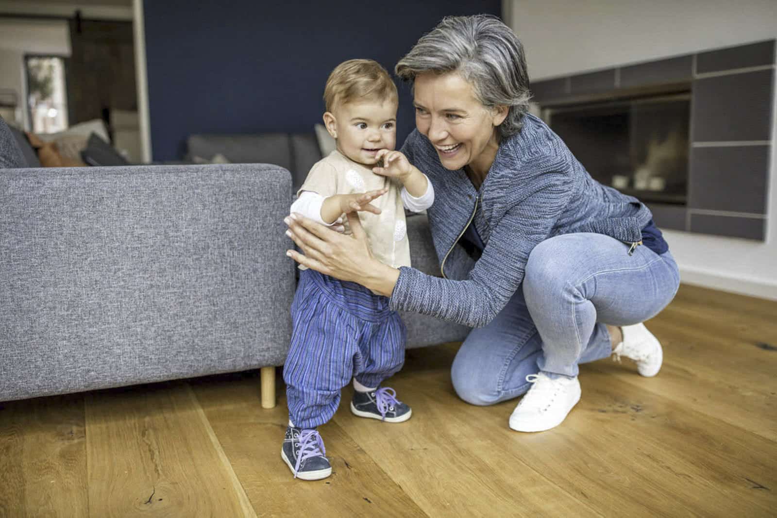 Eine lächelnde Frau hockt neben einem fröhlichen Kleinkind in einer blau gestreiften Hose, das sich in einem modernen Wohnzimmer mit Holzboden an einem grauen Sofa festhält. © Fotografie Tomas Rodriguez
